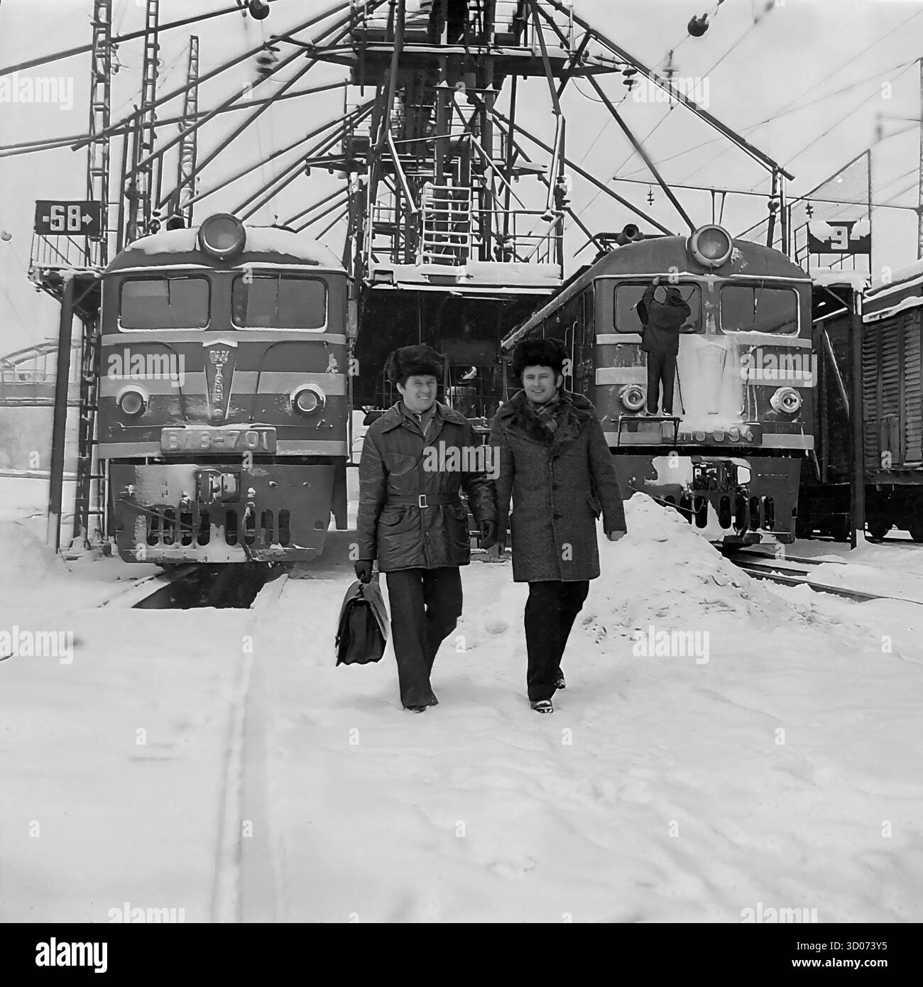 Deux cheminots soviétiques, dont un machiniste et vétéran (à droite), traversent un dépôt de chemin de fer enneigé à Sloviansk, dans les années 1970-1980 Archive photo en noir et blanc, URSS, RSS d'Ukraine. Les hommes, portant des manteaux d'hiver et des chapeaux de fourrure, marchent et parlent, avec un portant une mallette. En arrière-plan, on trouve des locomotives électriques soviétiques, dont un VL8 (ВЛ8-701), et une infrastructure ferroviaire complexe. Cette image montre la vie industrielle soviétique quotidienne, la profession de transport et les travailleurs dans des conditions hivernales difficiles. Un document précieux de la vie paisible dans le Donbass avant la guerre. Banque D'Images Deux cheminots soviétiques, dont un machiniste et vétéran (à droite), traversent un dépôt de chemin de fer enneigé à Sloviansk, dans les années 1970-1980 Archive photo en noir et blanc, URSS, RSS d'Ukraine. Les hommes, portant des manteaux d'hiver et des chapeaux de fourrure, marchent et parlent, avec un portant une mallette. En arrière-plan, on trouve des locomotives électriques soviétiques, dont un VL8 (ВЛ8-701), et une infrastructure ferroviaire complexe. Cette image montre la vie industrielle soviétique quotidienne, la profession de transport et les travailleurs dans des conditions hivernales difficiles. Un document précieux de la vie paisible dans le Donbass avant la guerre. Banque D'Images