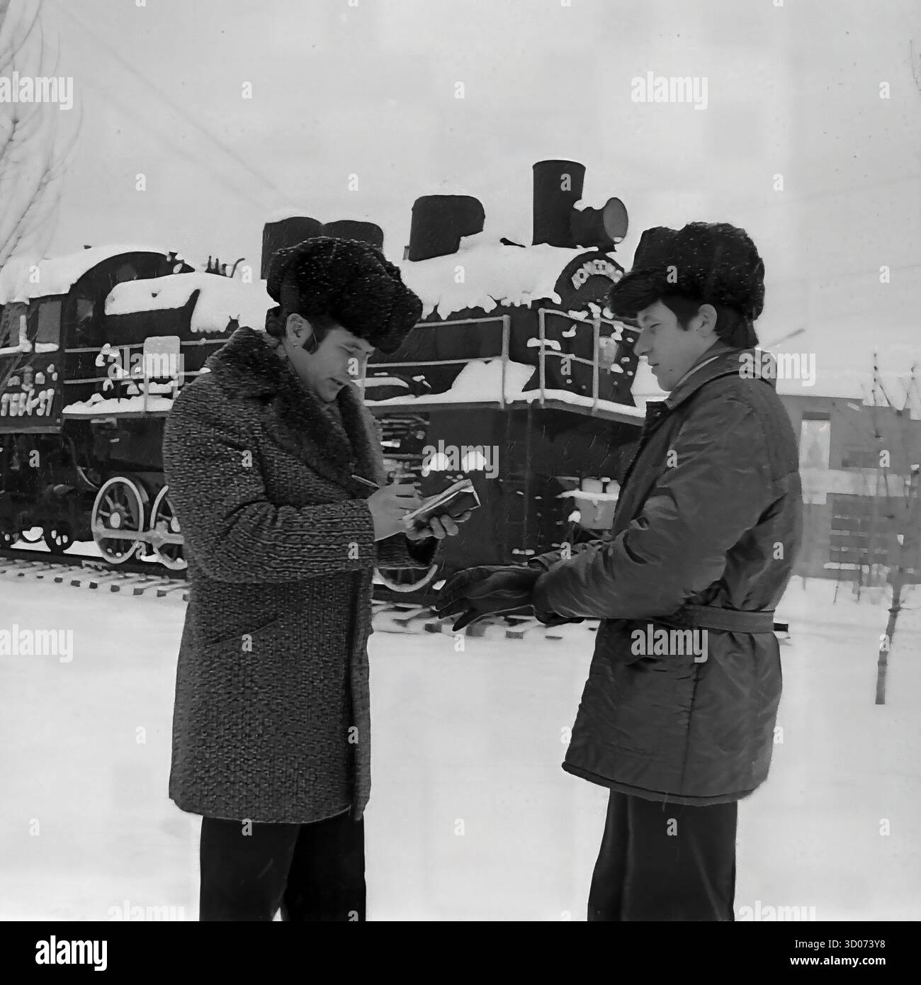 Deux cheminots soviétiques, un machiniste et vétéran (à gauche) et son collègue, se tiennent dans la neige à Sloviansk, dans les années 1970-1980 Archive photo en noir et blanc, URSS, RSS d'Ukraine. Ils ont une discussion devant un monument de locomotive à vapeur enneigé (modèle TE-64-37, 'Komsomolets'). Le vétéran écrit dans un cahier. Les deux hommes portent des manteaux d'hiver et des chapeaux de fourrure ('papakha'). Cette image montre la vie industrielle soviétique quotidienne, l'histoire des chemins de fer et un hiver rigoureux. Un document précieux de la vie paisible dans le Donbass avant la guerre. Banque D'Images Deux cheminots soviétiques, un machiniste et vétéran (à gauche) et son collègue, se tiennent dans la neige à Sloviansk, dans les années 1970-1980 Archive photo en noir et blanc, URSS, RSS d'Ukraine. Ils ont une discussion devant un monument de locomotive à vapeur enneigé (modèle TE-64-37, 'Komsomolets'). Le vétéran écrit dans un cahier. Les deux hommes portent des manteaux d'hiver et des chapeaux de fourrure ('papakha'). Cette image montre la vie industrielle soviétique quotidienne, l'histoire des chemins de fer et un hiver rigoureux. Un document précieux de la vie paisible dans le Donbass avant la guerre. Banque D'Images