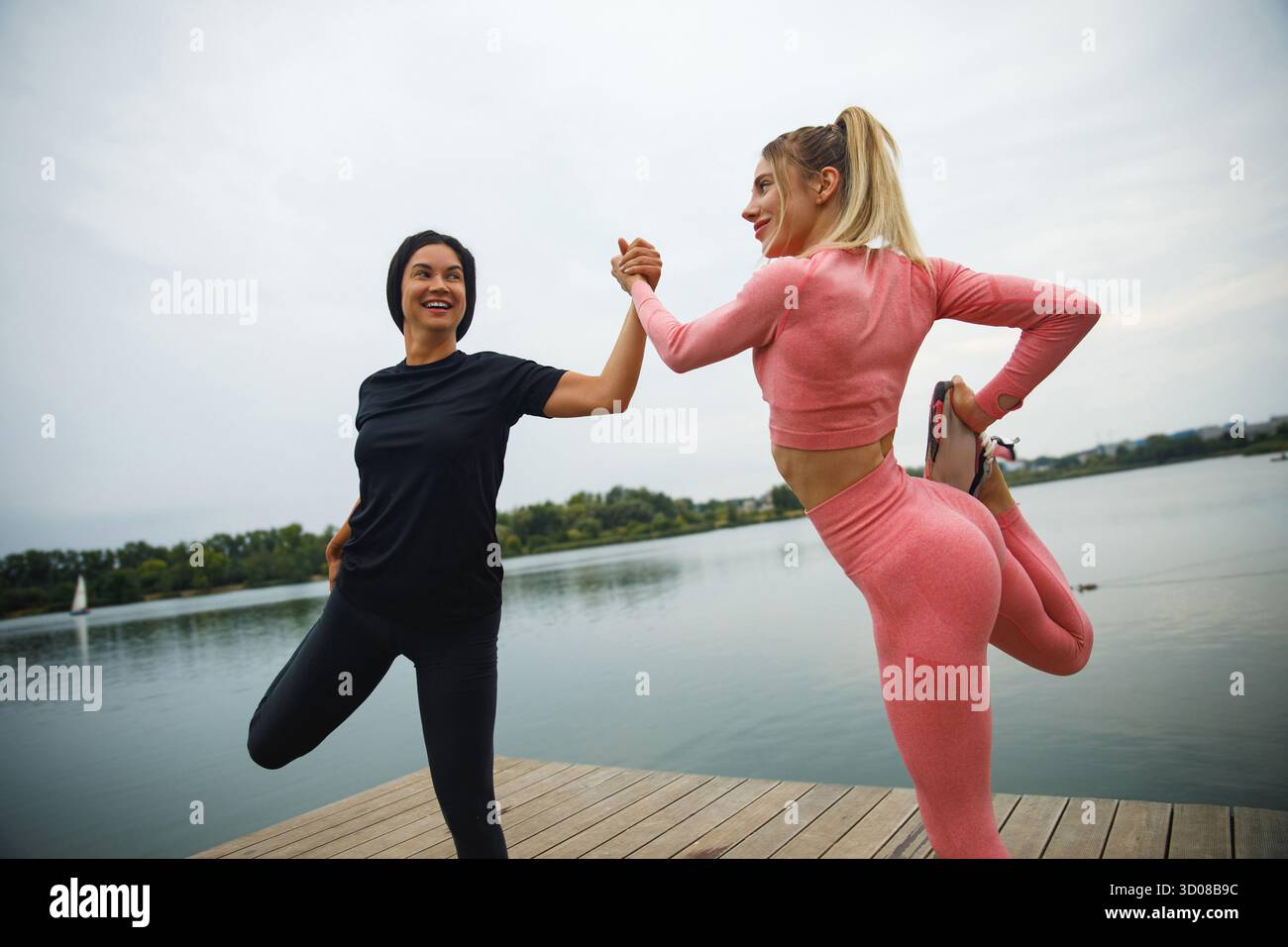 Deux femmes s’engagent dans une séance de yoga amusante au bord de l’eau, riant et se soutenant mutuellement. Leur énergie joyeuse renforce l'atmosphère paisible du o Banque D'Images