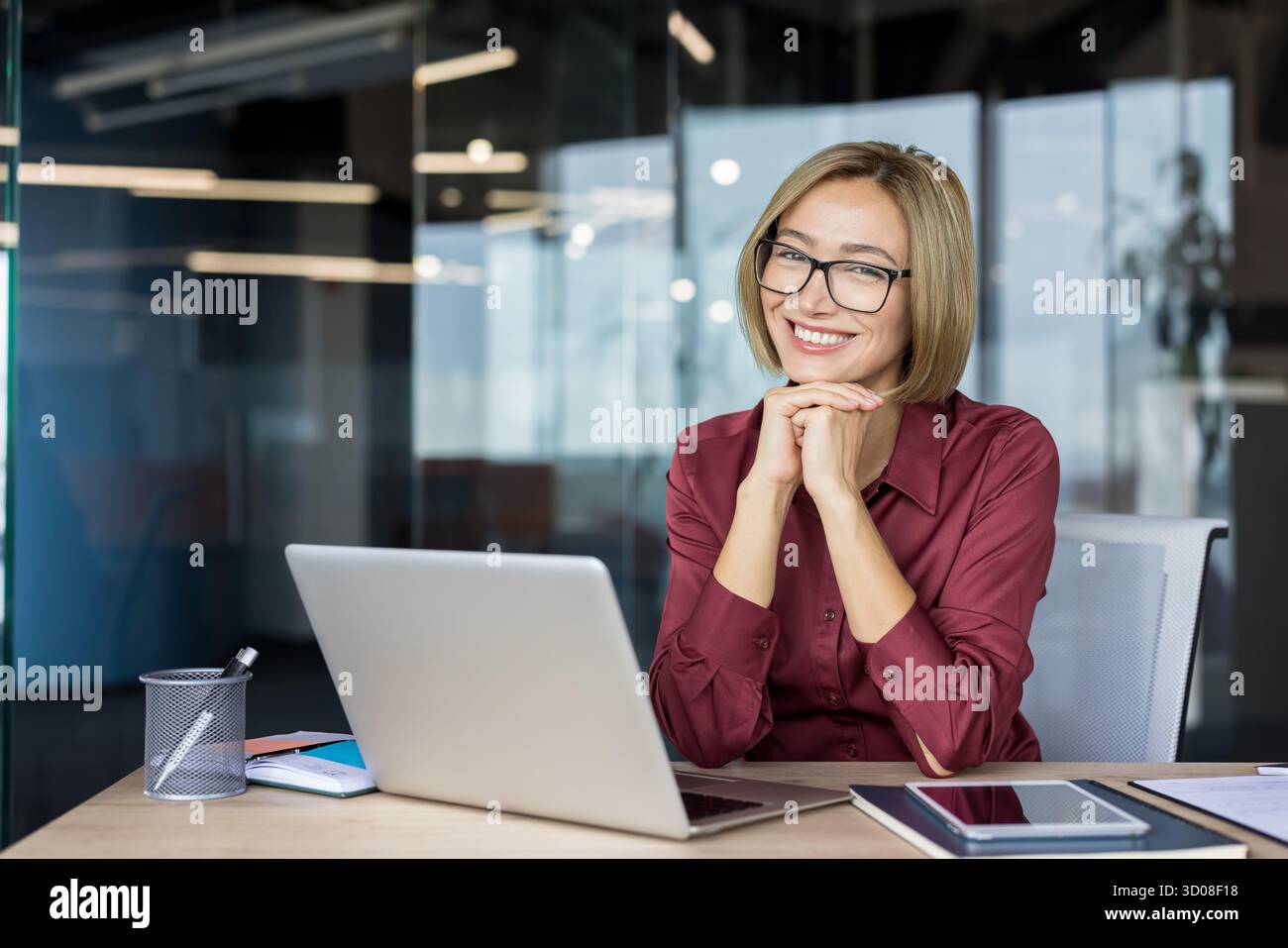Femme d'affaires professionnelle souriant avec confiance, se reposant le menton sur les mains au bureau avec ordinateur portable dans le bureau moderne, lunettes, portrait détendu et concentré transmettant le succès et le leadership Banque D'Images