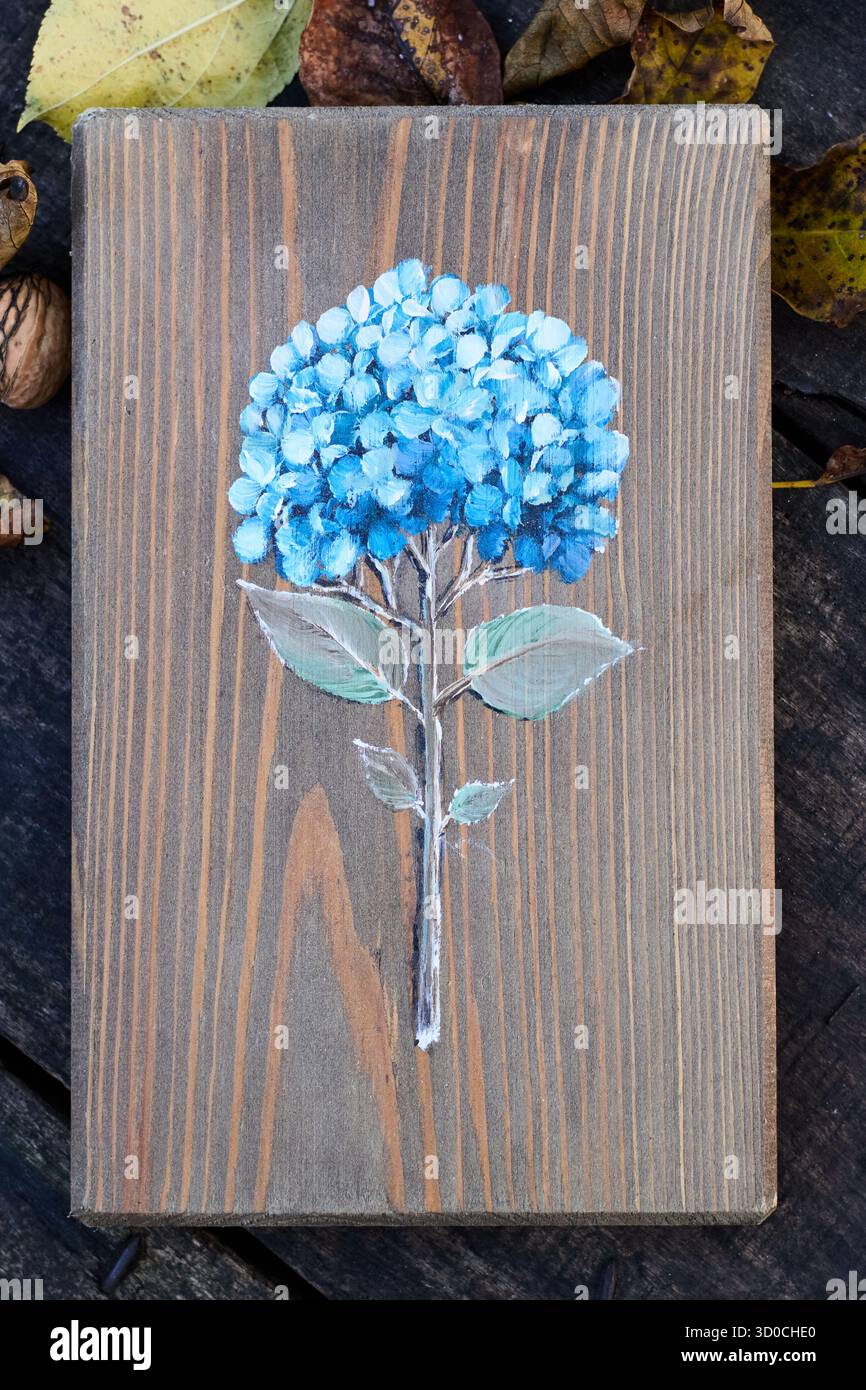 Hortensia bleue peinte à la main sur une planche de bois brun, entourée de feuilles sèches et de noix sur une table en bois rustique, décoration florale artistique Banque D'Images