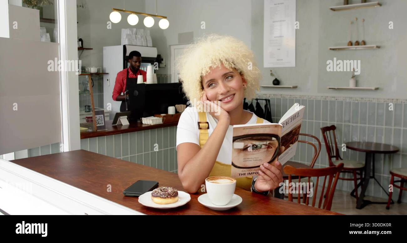 Femme de lecture portant une robe jaune penchée la tête sur la main au comptoir du café, avec livre et cappuccino Banque D'Images
