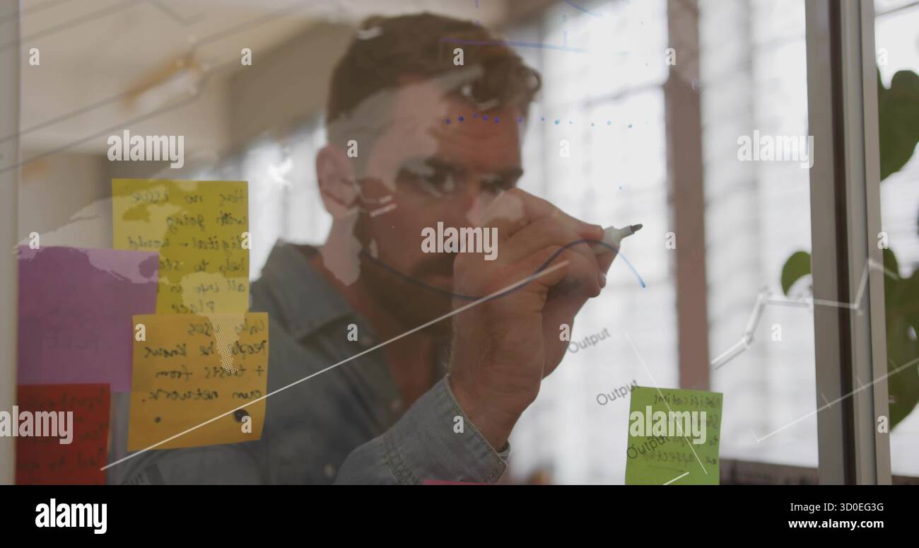 Homme senior dessinant une courbe montante sur un tableau transparent au bureau, avec des notes adhésives colorées et un marqueur Banque D'Images