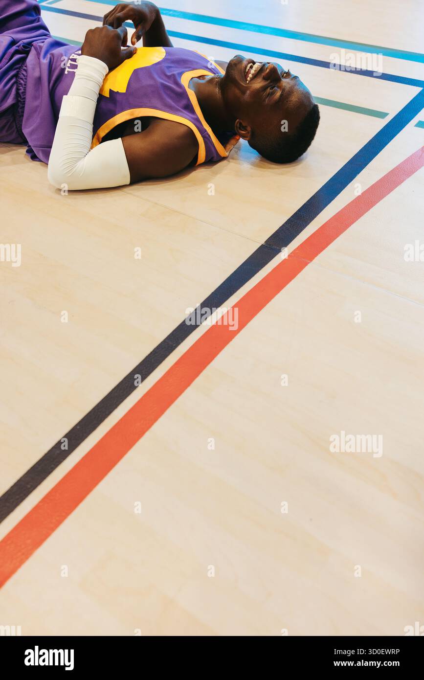 Joueur de basket épuisé allongé sur le terrain et riant avec soulagement après un match intense, symbolisant la persévérance, la joie et la libération émotionnelle i. Banque D'Images