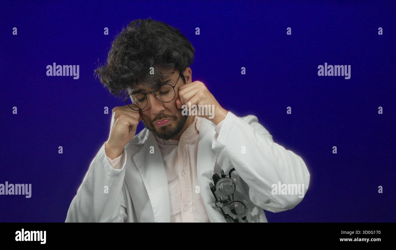 Jeune homme en blouse de laboratoire blanche avec des lunettes prétendant pleurer sur un fond bleu, mettant en valeur un geste humoristique et expressif. Banque D'Images