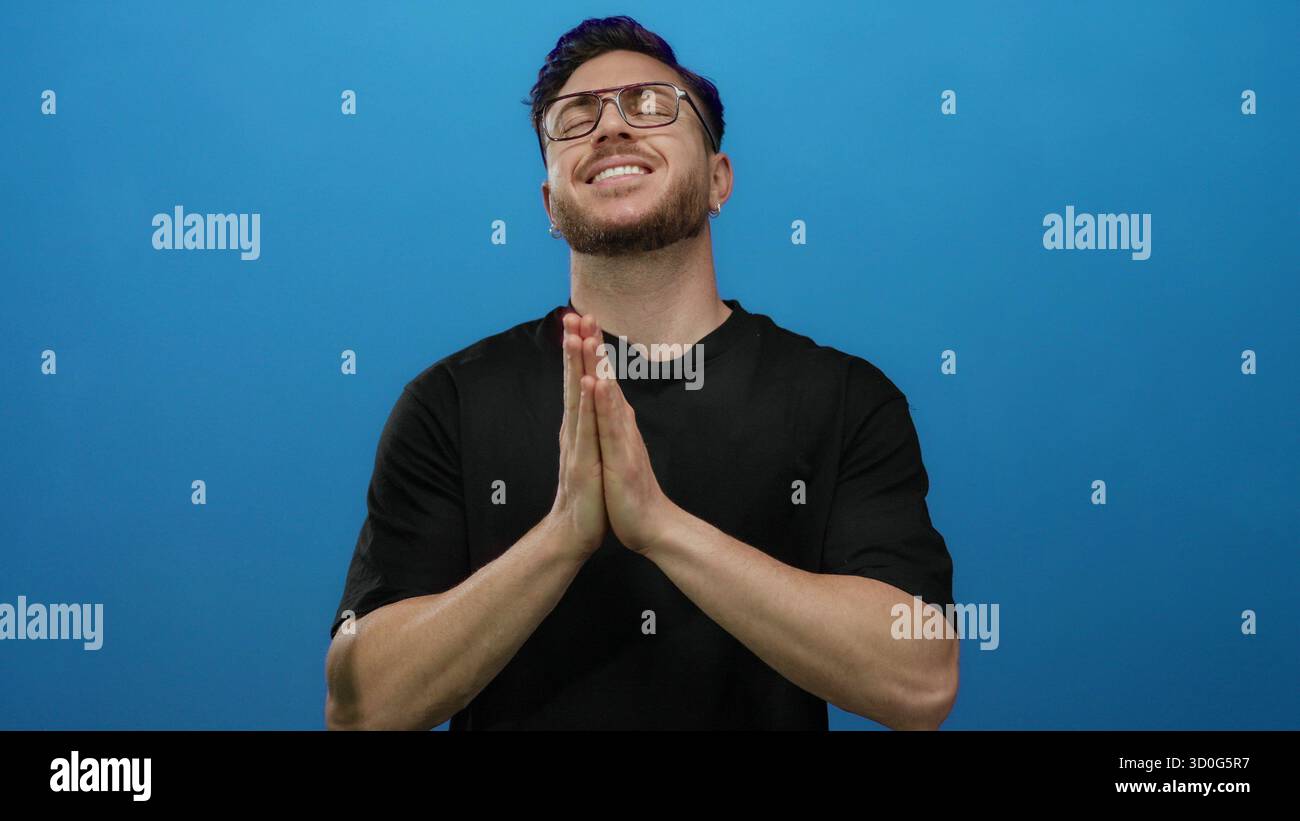 Jeune homme à la barbe priant les yeux fermés sur fond bleu, exprimant espoir et spiritualité dans un cadre isolé. Banque D'Images
