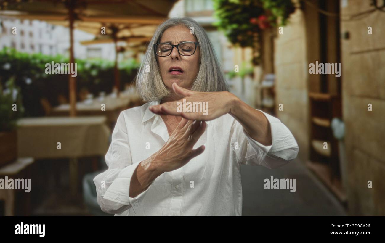 Femme avec des cheveux gris et des lunettes faisant le geste de la main Time Out sur la terrasse du restaurant à l'extérieur, chemise blanche avec col ouvert, mains levées ; limite con Banque D'Images
