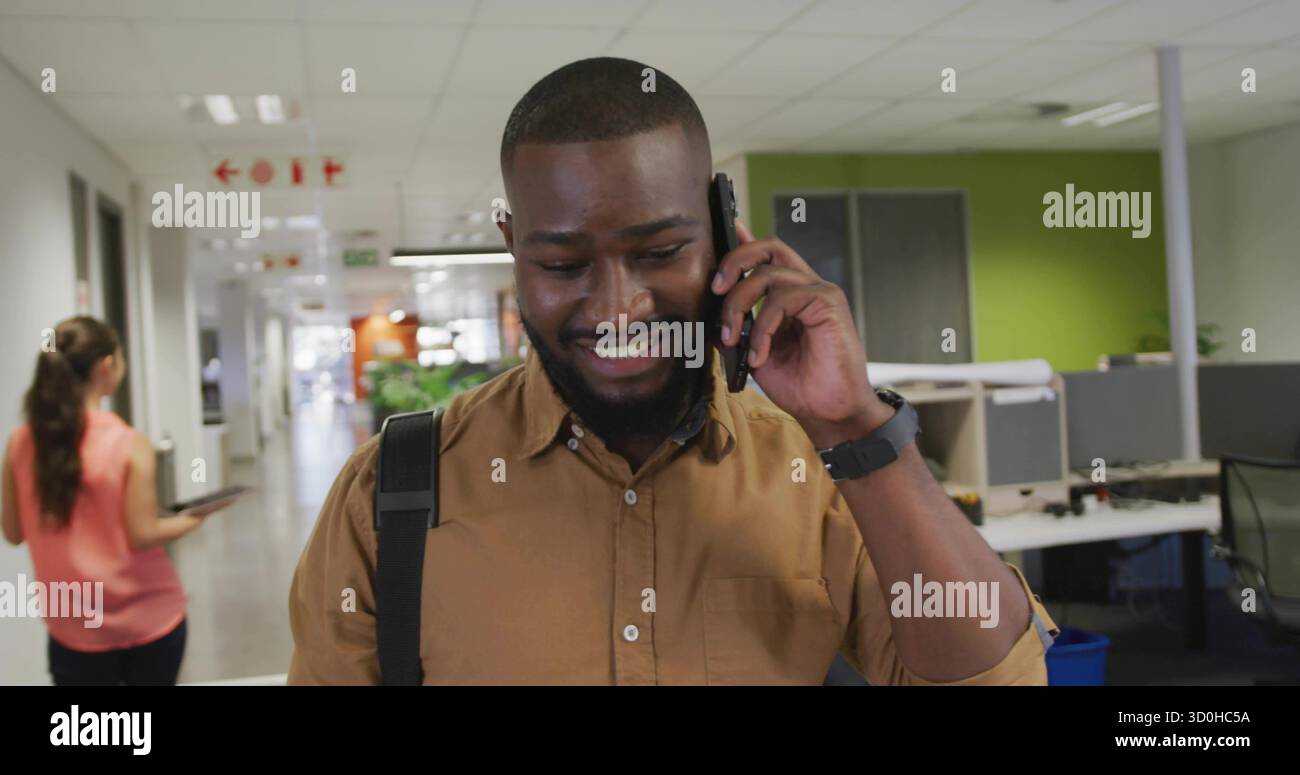 Travailleur marchant en chemise brune parlant sur smartphone à travers le couloir avec sac à dos, plante en pot. Professionnel, corporatif, moderne, collaboratif, conne Banque D'Images