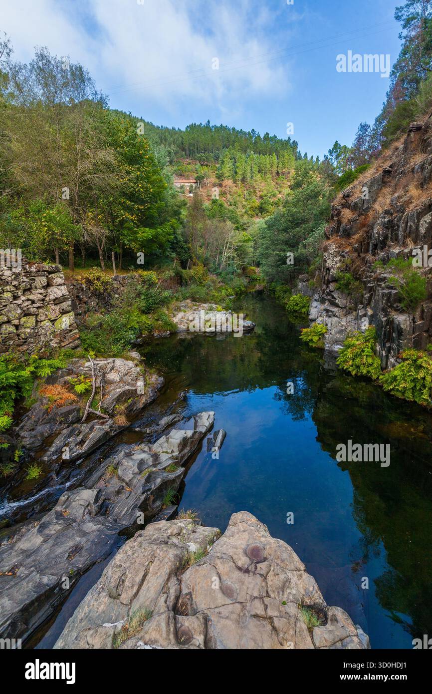 Une petite cascade coule sur des rochers sombres et superposés dans une rivière ruisselante, encadrée par une forêt verdoyante et des falaises escarpées sous un ciel couvert Banque D'Images