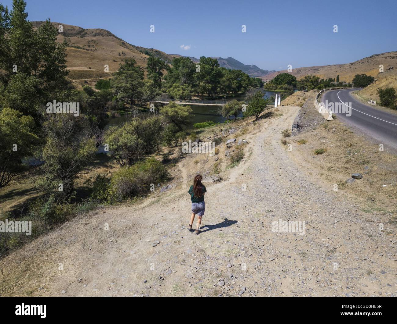 Vue aérienne d'une figure solitaire debout sur un chemin poussiéreux surplombant une rivière bordée d'arbres luxuriants à côté d'une route sinueuse, Samtskhe-Javakheti, Samtskhe-Javakheti, Géorgie. Banque D'Images