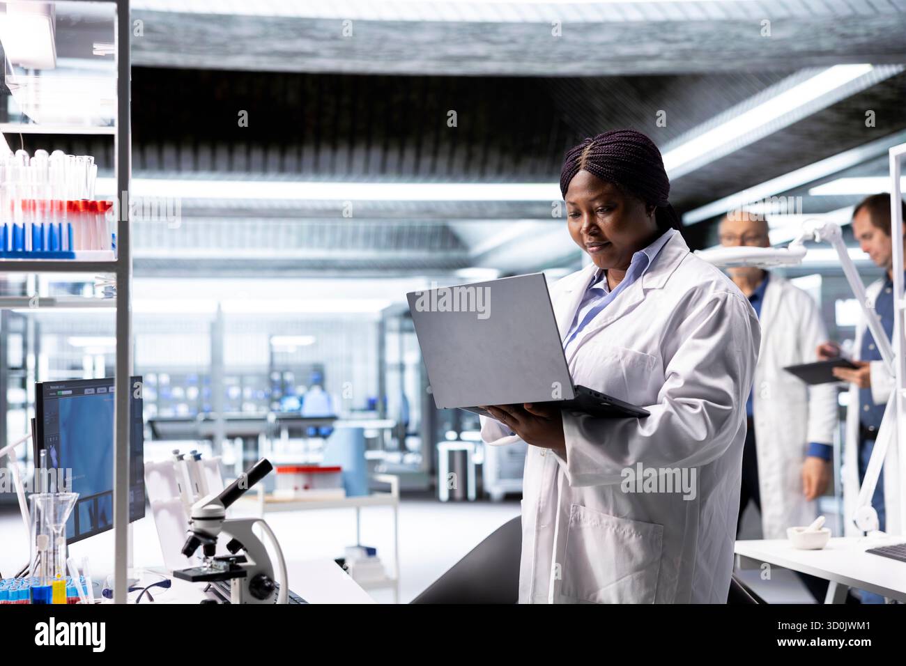 Afro-américaine scientifique travaille dans un laboratoire moderne, menant des expériences avec des équipements de pointe et un ordinateur portable. Axé sur la biologie moléculaire, la biotechnologie et l'innovation dans le domaine de la santé. Banque D'Images