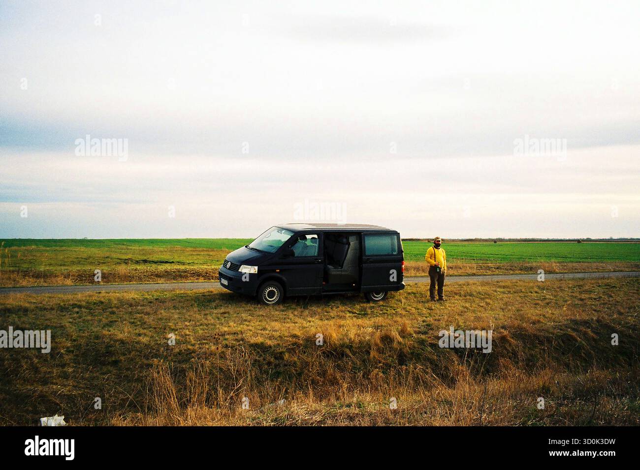 Homme dans un pull jaune debout à côté d'une VW T5 stationnée, regardant dans la vaste plaine ouverte derrière lui, capturée dans des tons analogiques chauds. Banque D'Images