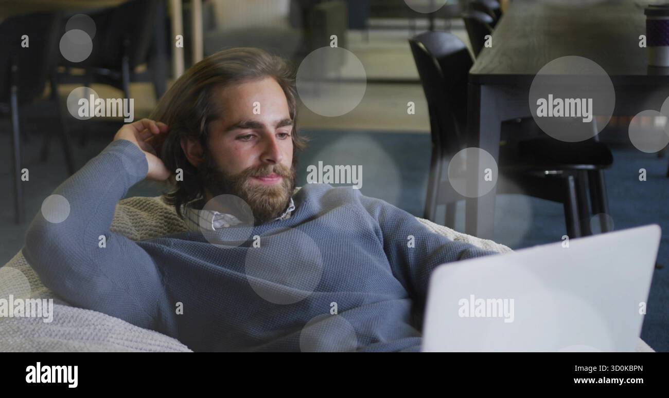 Homme barbu inclinable naviguant sur l'ordinateur portable dans une chaise longue dans le salon du bureau, avec tasse à café et chaises Banque D'Images