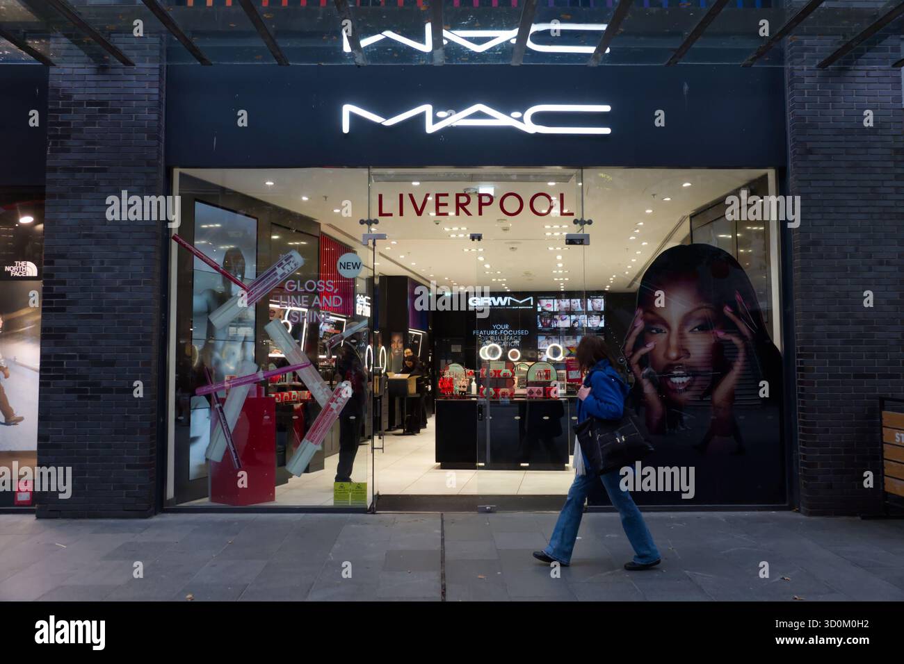 Photo d'une femme passant devant le magasin MAC Cosmetics illuminé de Liverpool One, avec des affiches lumineuses et des présentoirs de maquillage à l'intérieur. Banque D'Images