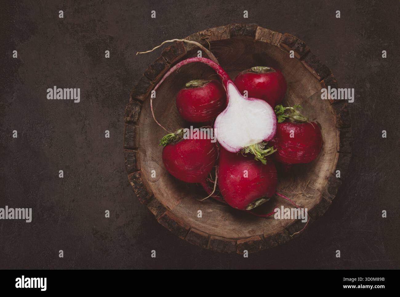 Une collection de radis rouges vibrants se trouve dans un bol en bois sculpté sur une surface sombre et texturée. Les légumes frais sont parfaits pour les salades ou la garnishe Banque D'Images
