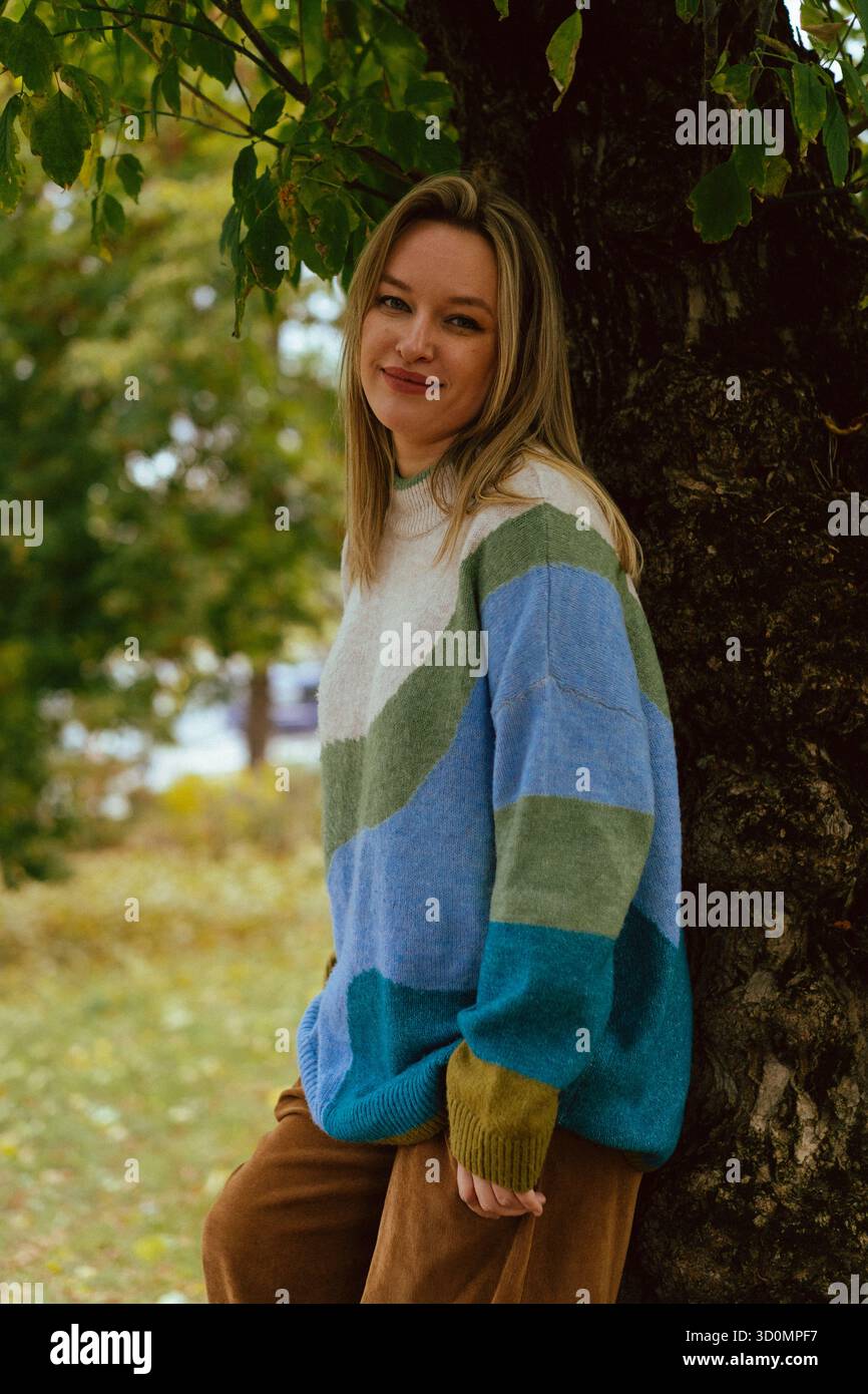 Jeune femme souriante appuyée contre un tronc d'arbre dans un pull coloré Banque D'Images