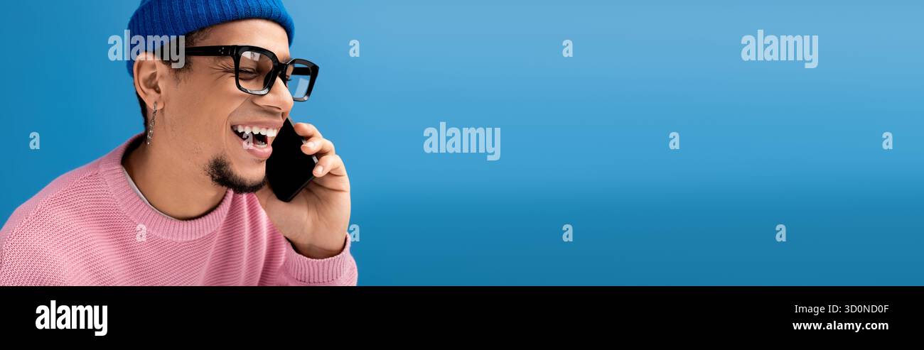 Beau jeune homme vêtu de vêtements à la mode discute joyeusement au téléphone dans un contexte dynamique. Banque D'Images