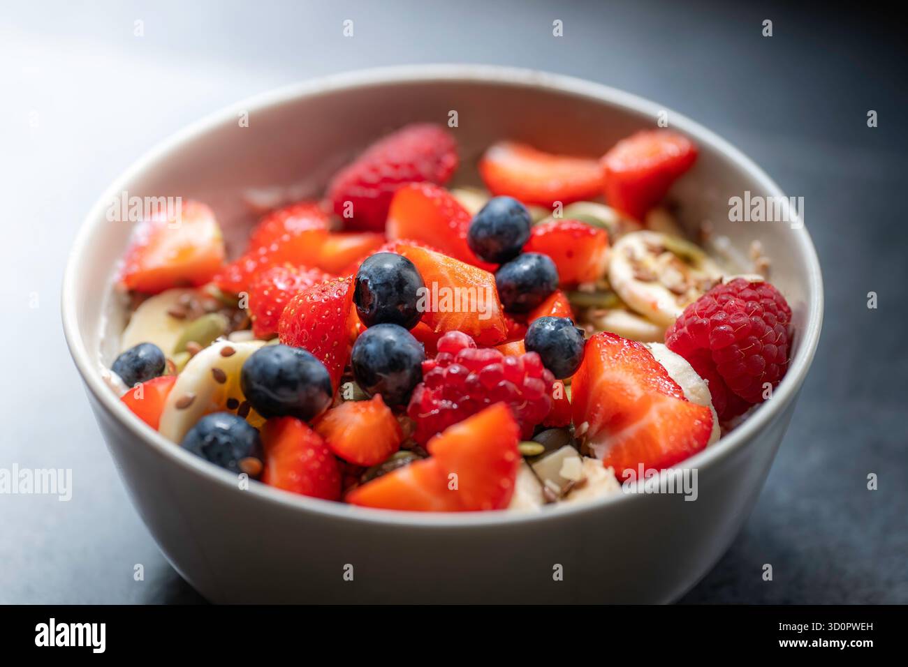 Flocons d'avoine avec des fruits dans un bol beige sur un comptoir anthracite. Le bol est rempli de fraises, de bleuets, de framboises, de bananes et de graines de lin Banque D'Images