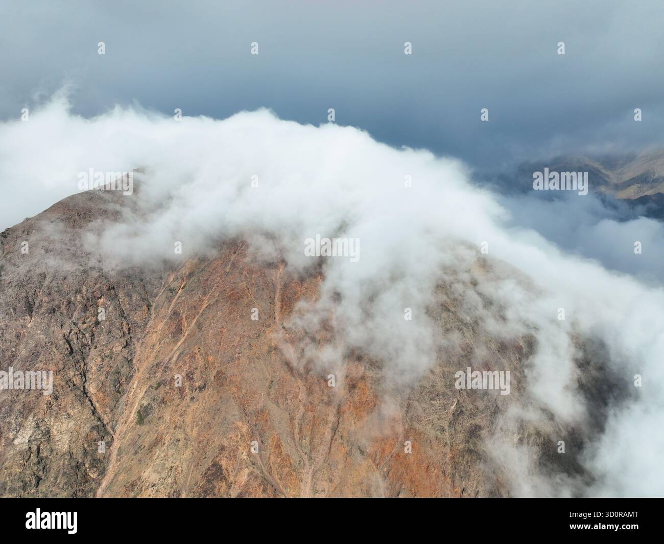 Gros plan aérien extrême du sommet accidenté, texturé et brun-rougeâtre d'une montagne avec une épaisse couche de nuages blancs roulant et se répandant sur le sommet Banque D'Images