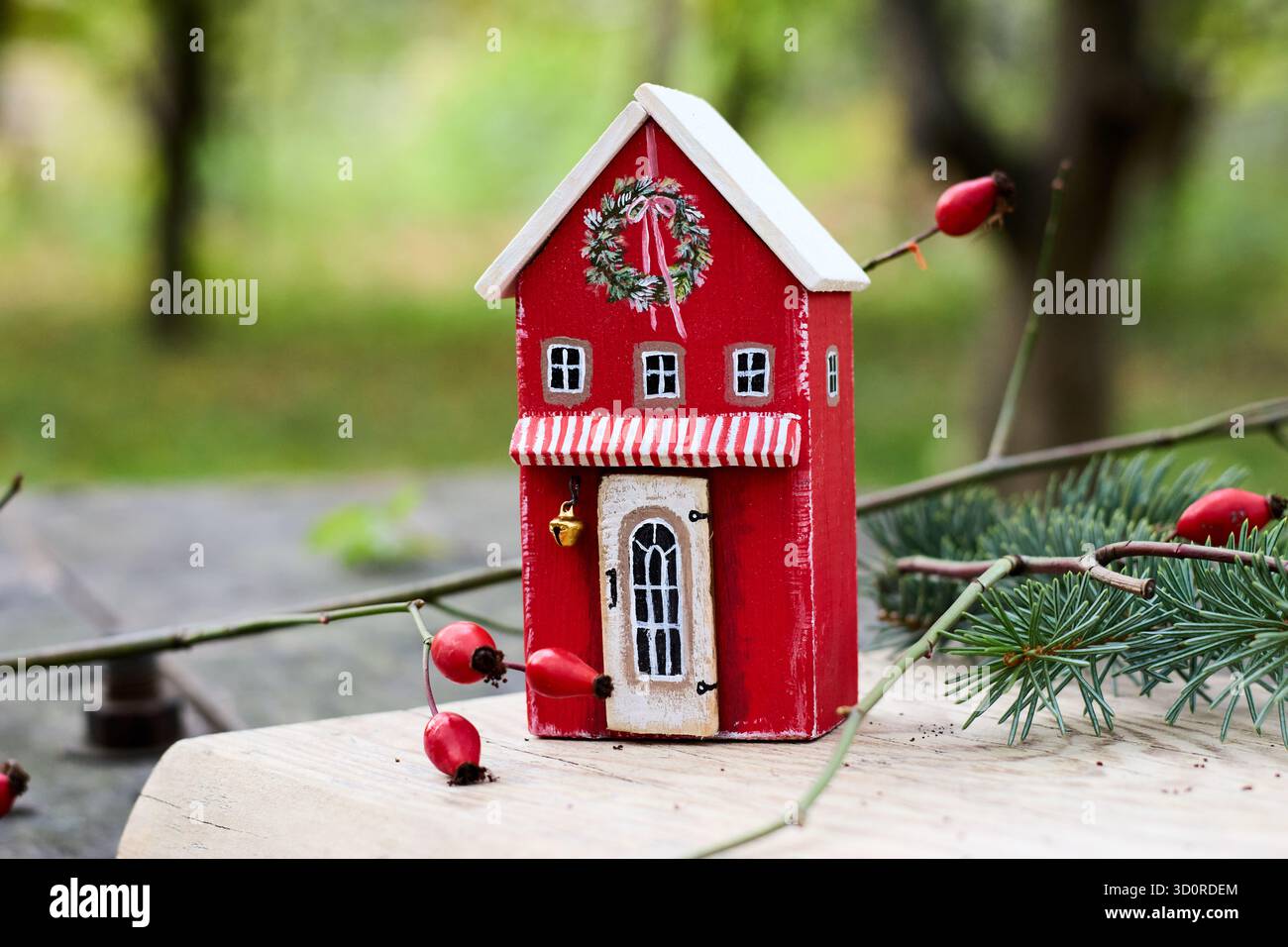 Une charmante maison de jouet en bois rouge ornée d'une couronne festive, évoquant l'esprit de Noël et le charme rustique sur un fond flou. Banque D'Images