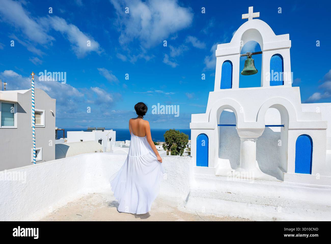 Une jolie femme brune dans une longue robe blanche surplombe la mer depuis un clocher blanc dans la ville blanchie à la chaux de colline d'Oia, Santorin Grèce Banque D'Images