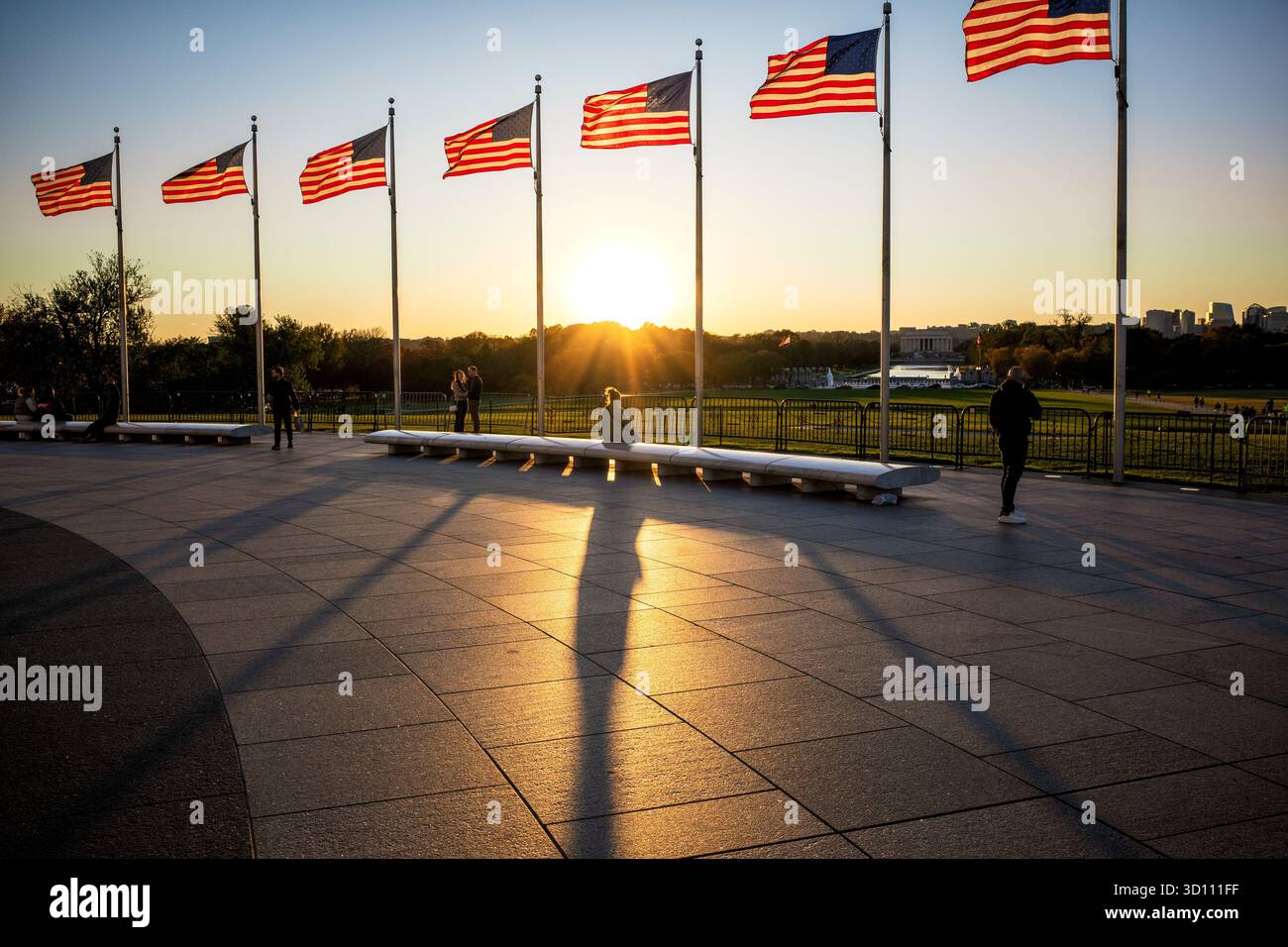 Drapeaux américains au coucher du soleil Washington DC // WASHINGTON DC — les drapeaux américains sont baignés de lumière dorée juste avant le coucher du soleil sur le National Mall. Le soleil bas projette de longues ombres sur la zone pavée au premier plan, créant un effet dramatique. En arrière-plan, le Lincoln Memorial Reflecting Pool et le Lincoln Memorial sont des monuments emblématiques visibles de la capitale nationale. Cette zone est un site important pour les monuments nationaux et les rassemblements publics. Banque D'Images