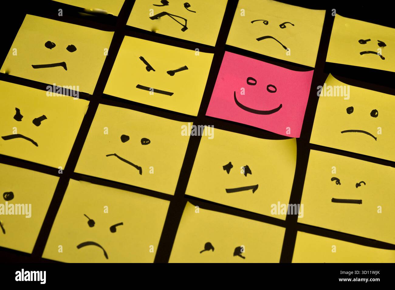 15 papiers jaunes placés parallèlement les uns aux autres avec des visages dessinés sur eux exprimant la colère ou la tristesse, face à un papier rose avec un visage exprimant le happin Banque D'Images