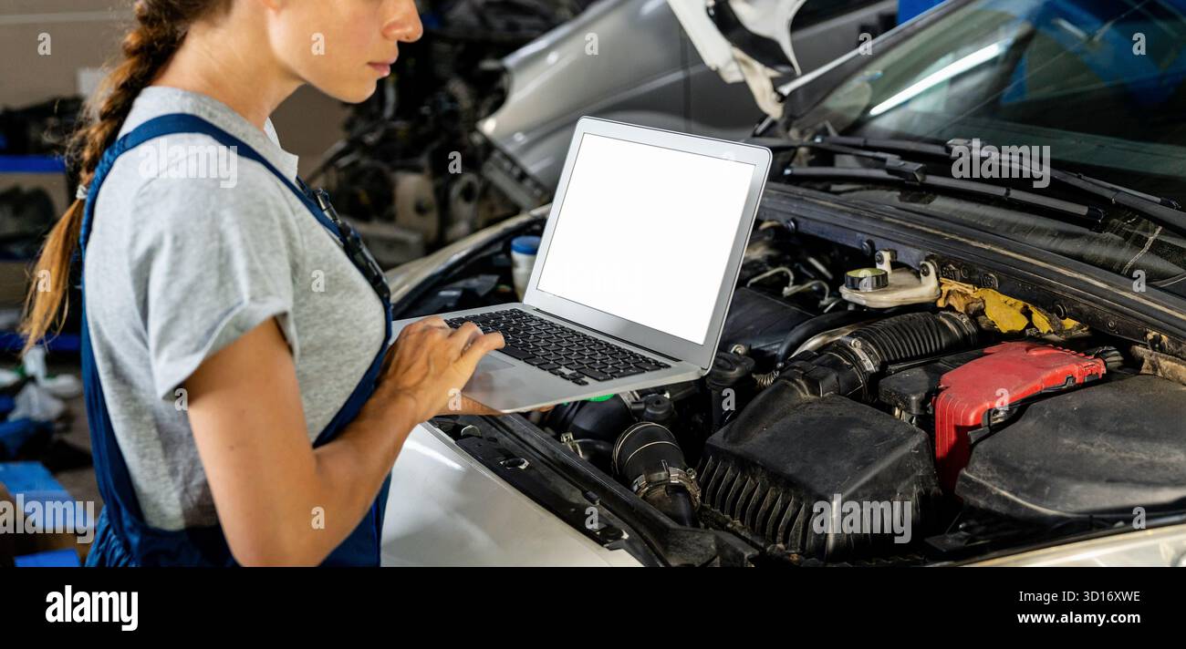 Ordinateur portable avec écran blanc dans les mains du mécanicien automobile féminin au-dessus du capot de voiture ouvert Banque D'Images