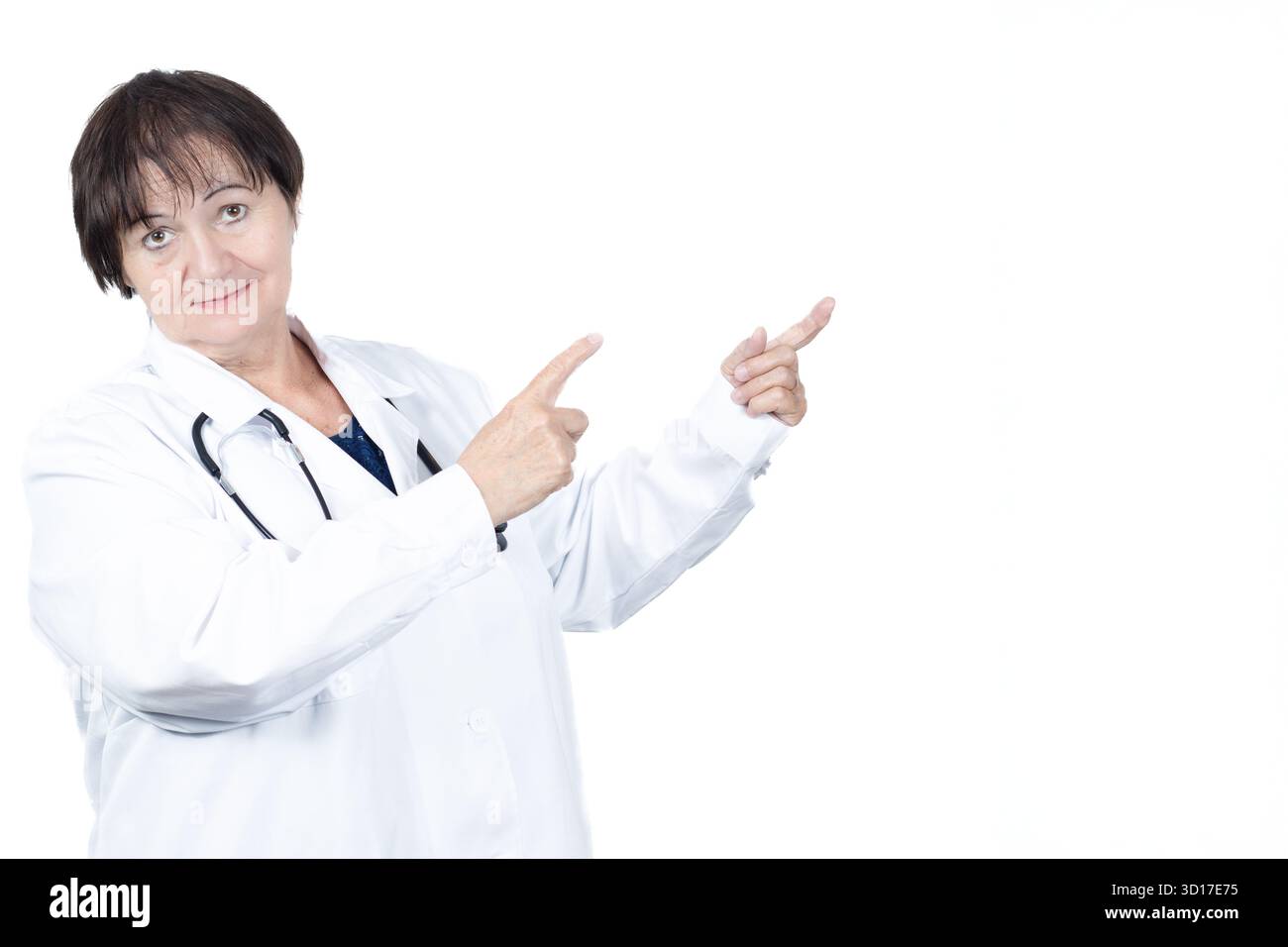 Médecin femme expérimentée portant une blouse de laboratoire et un stéthoscope, souriant tout en pointant deux doigts vers l'espace vide sur un fond blanc, suggérant Banque D'Images