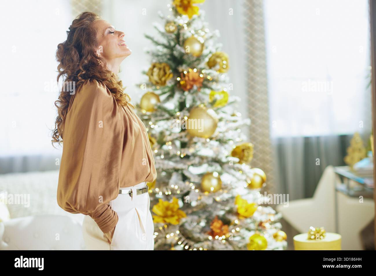 Femme aux cheveux bruns bouclés, les yeux fermés, se tient devant l'arbre de Noël décoré. Une scène intérieure très éclairée évoque une joie et une tranquillité festives profondes Banque D'Images