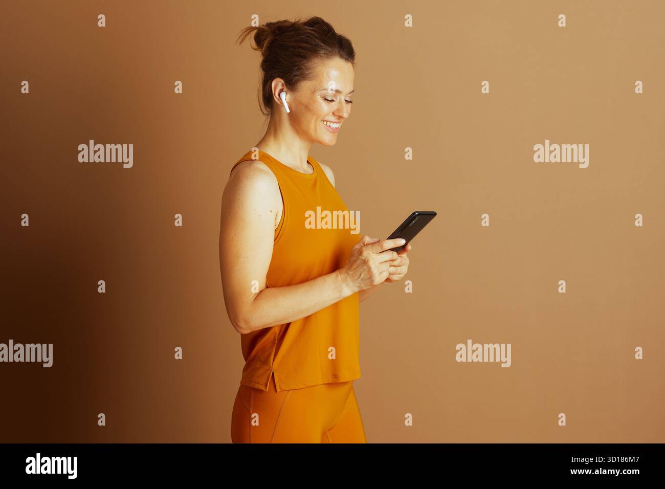 Une femme d'âge moyen souriante portant des vêtements de sport jaune moutarde et des écouteurs est vue de profil, regardant attentivement son smartphone. Elle incarne le e numérique Banque D'Images