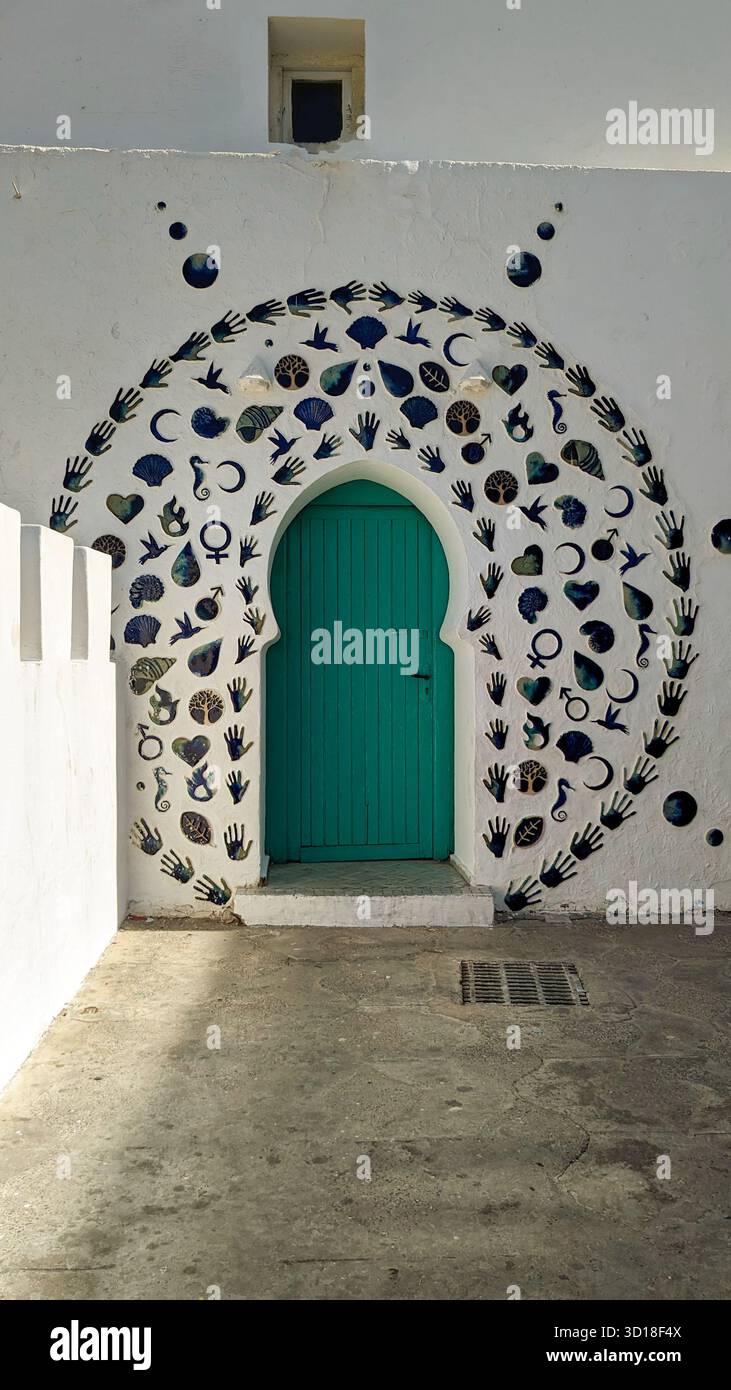 Porte arquée turquoise encadrée par une mosaïque artistique de mains, de coquillages, de coeurs et de symboles culturels sur un mur texturé blanc - Image de stock capturée avec un smartphone