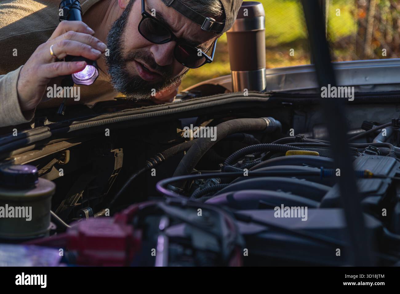 Un homme se penche au-dessus d'un capot de voiture ouvert, en utilisant une lampe de poche pour examiner le moteur de près. La lumière du soleil filtre à travers les arbres, créant une atmosphère chaleureuse. Banque D'Images