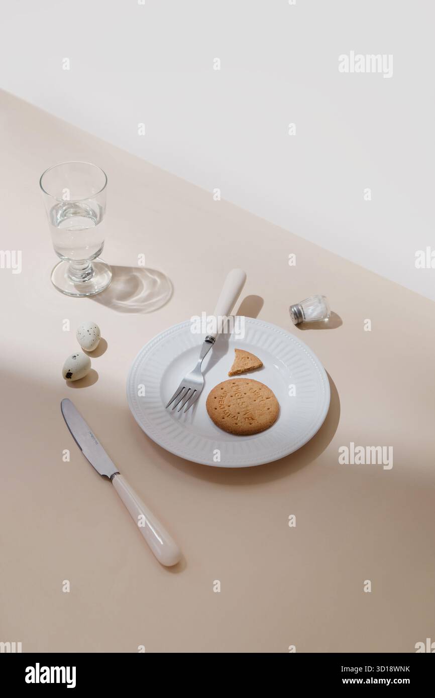 Biscuit sur assiette avec fourchette et verre d'eau. Cookie et fourchette sur plaque blanche avec verre d'eau, petit déjeuner minimaliste nature morte sur table beige Banque D'Images