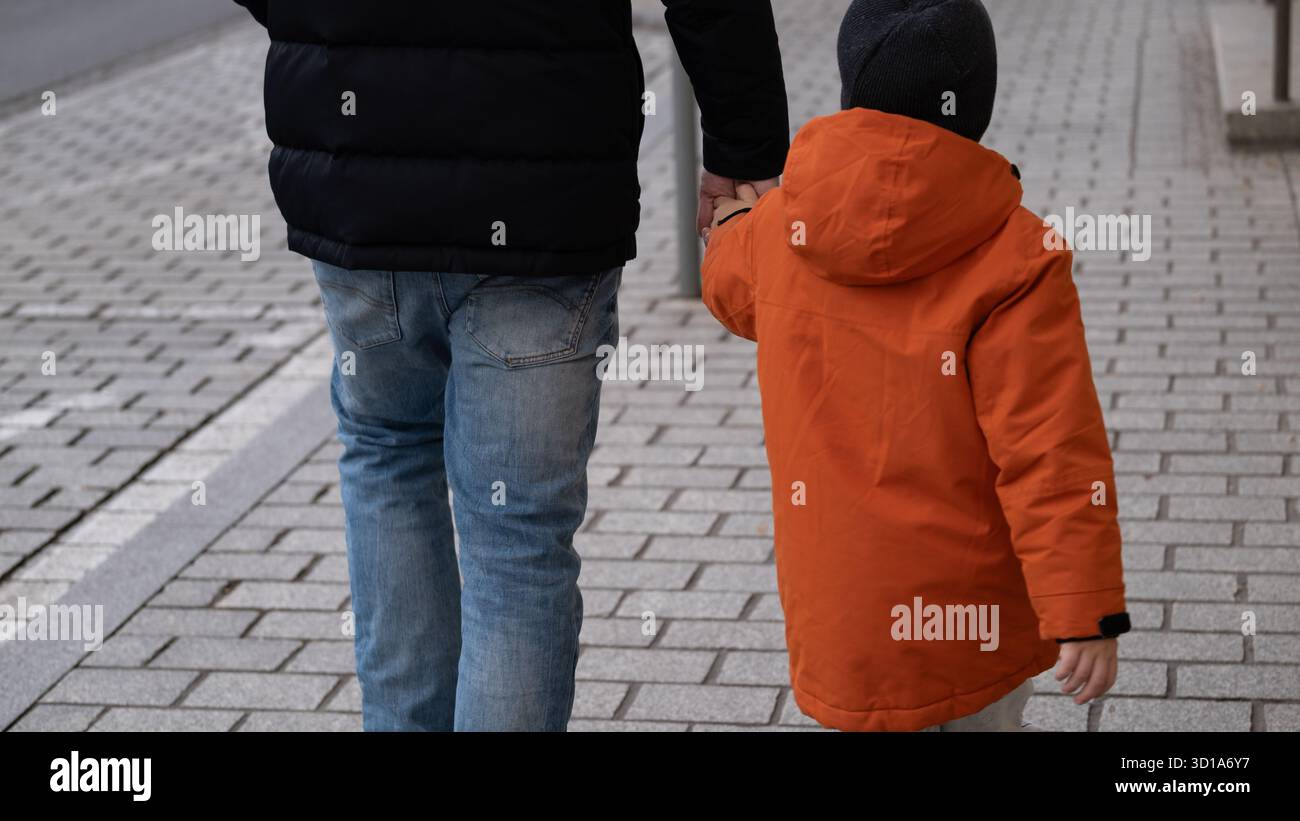 Vue arrière d'un homme et d'un enfant marchant ensemble sur un trottoir de la ville, se tenant par la main. L'enfant porte une veste et un chapeau orange, tandis que l'adulte est habillé Banque D'Images