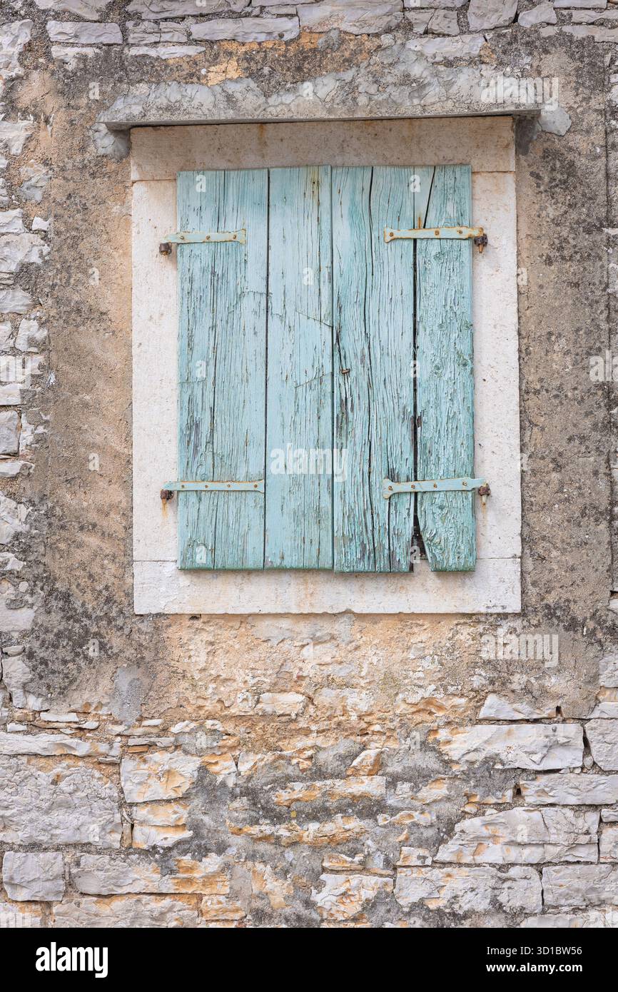 Un mur en pierre altérée encadre une inscription rouge fanée au-dessus d'un volet en bois bleu usé. La façade rustique évoque le patrimoine, les textures vieillissantes, et le calme, Banque D'Images