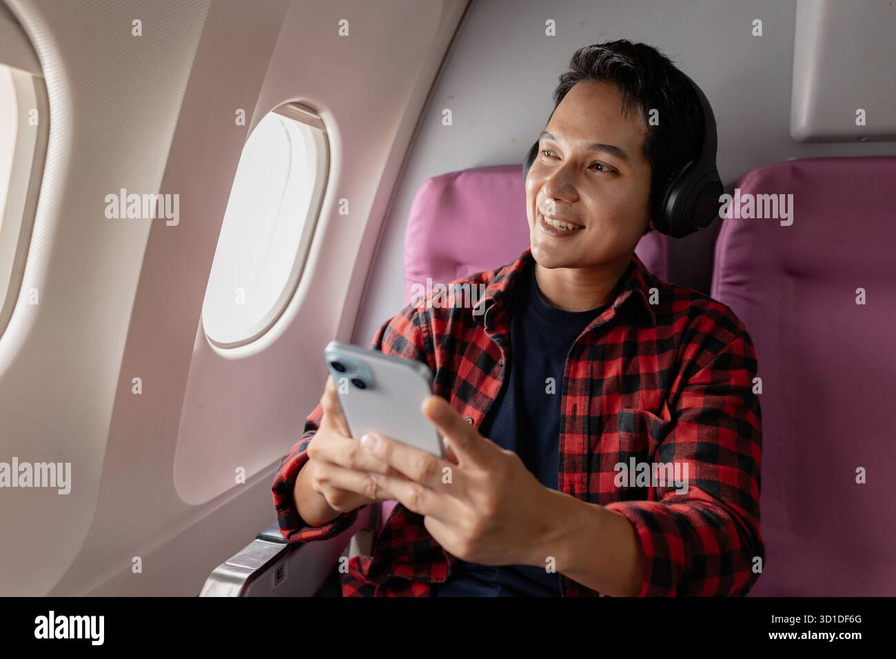 Un jeune homme joyeux aime son vol tout en utilisant son smartphone et en portant des écouteurs. Assis près de la fenêtre de l'avion, il montre de l'excitation et happ Banque D'Images