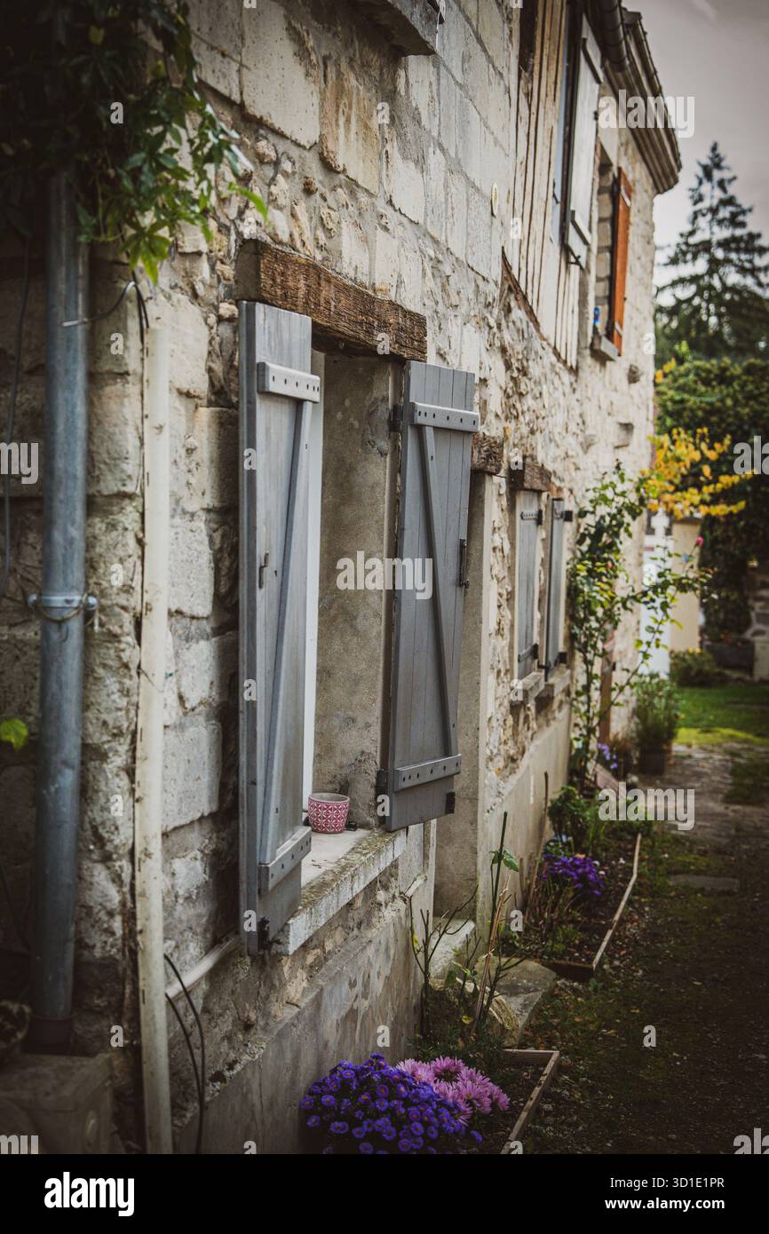 Un charmant bâtiment en pierre avec des volets gris altérés et un pot de fleurs vibrant ajoute une touche de couleur à la scène. Banque D'Images
