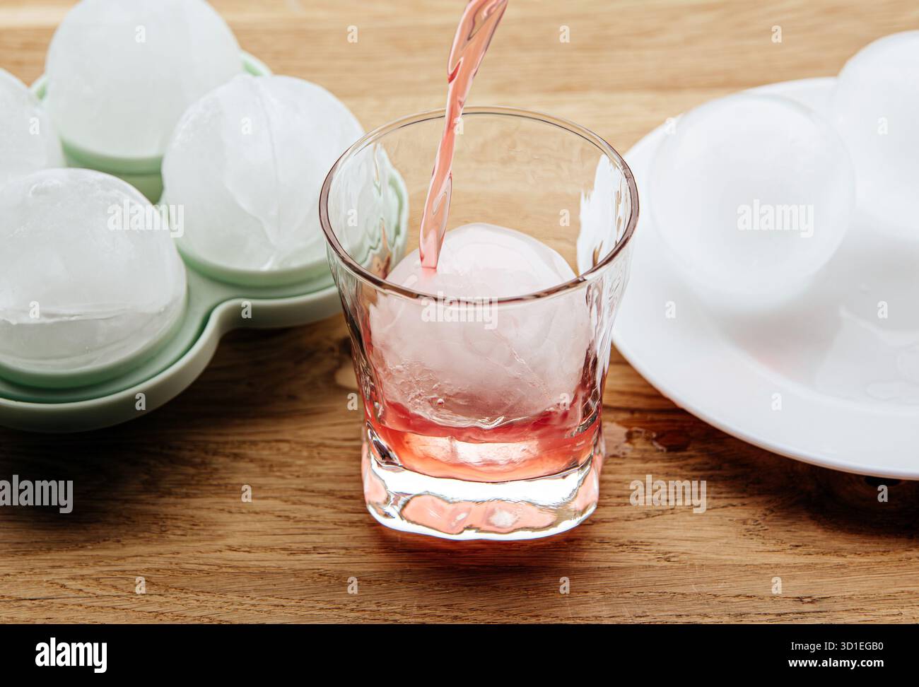 Un grand glaçon rond dans un verre, avec une boisson versée dessus. En arrière-plan, des glaçons ronds dans un moule en silicone sur une table de cuisine. Banque D'Images