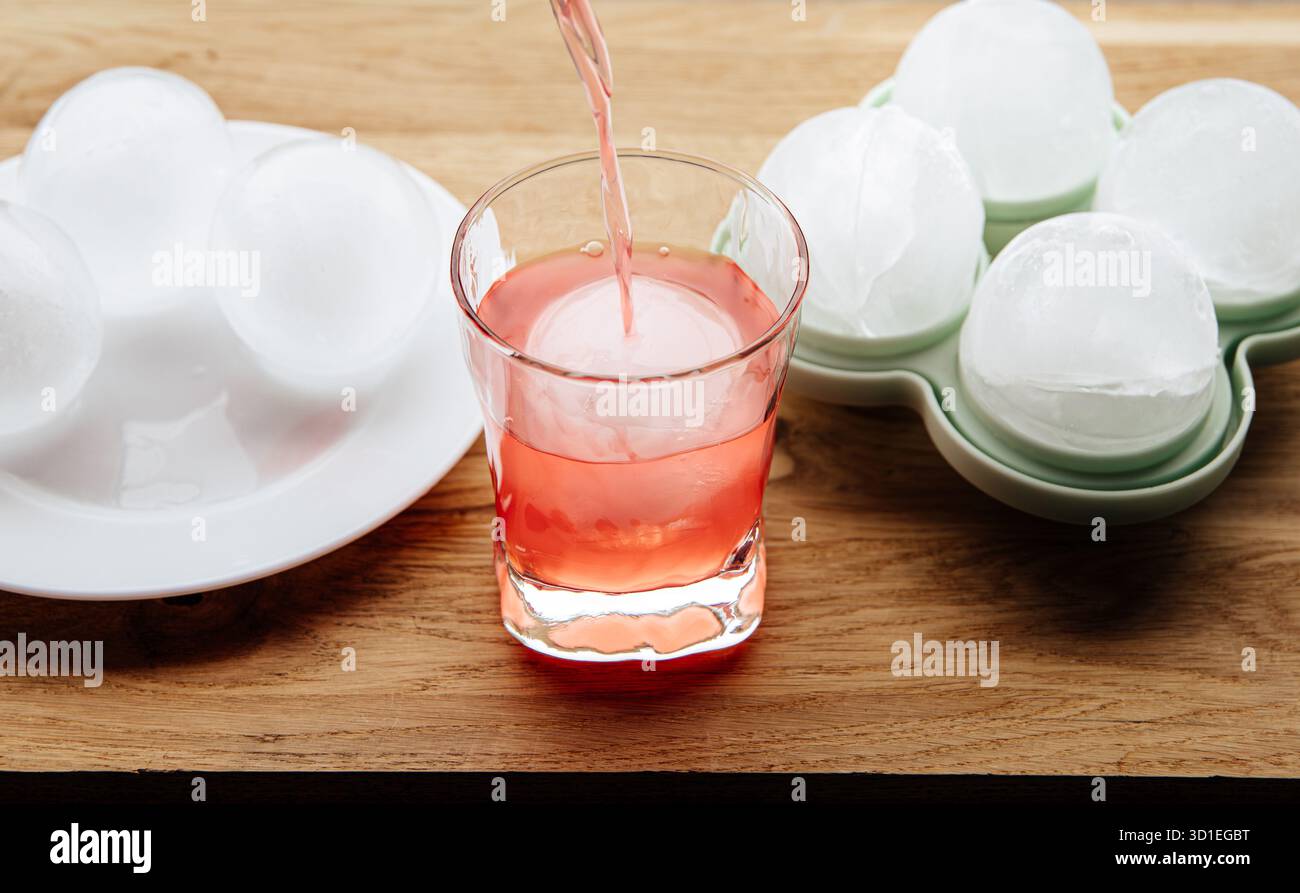 Un grand glaçon rond dans un verre, avec une boisson versée dessus. En arrière-plan, des glaçons ronds dans un moule en silicone sur une table de cuisine. Banque D'Images