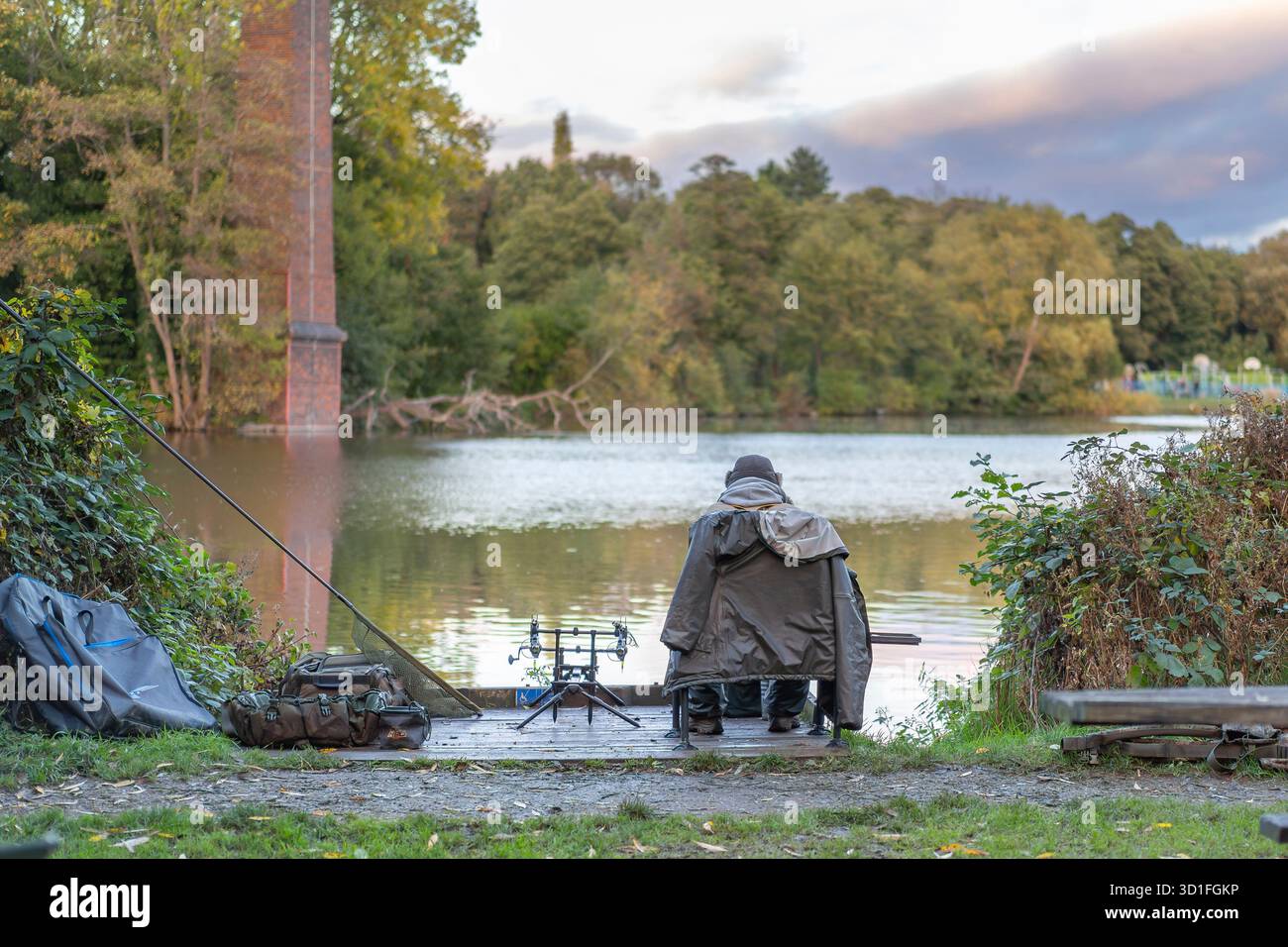 Vue arrière d'un pêcheur isolé / pêcheur assis avec une canne et tacle / équipement au bord d'une piscine britannique, manteau sur les épaules, pêche sur un jour d'automne frais. Banque D'Images