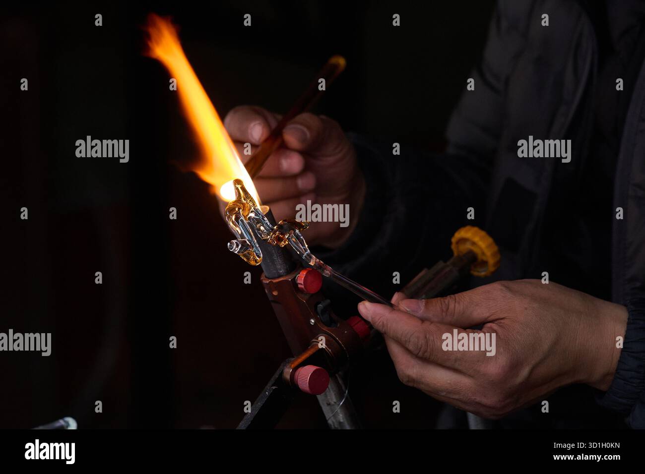 Artiste souffleur de verre démontrant la technique de la flamme de la torche, cadre d'atelier nocturne, personne dans la veste sombre actionnant la torche de verre, pratique artistique de Banque D'Images