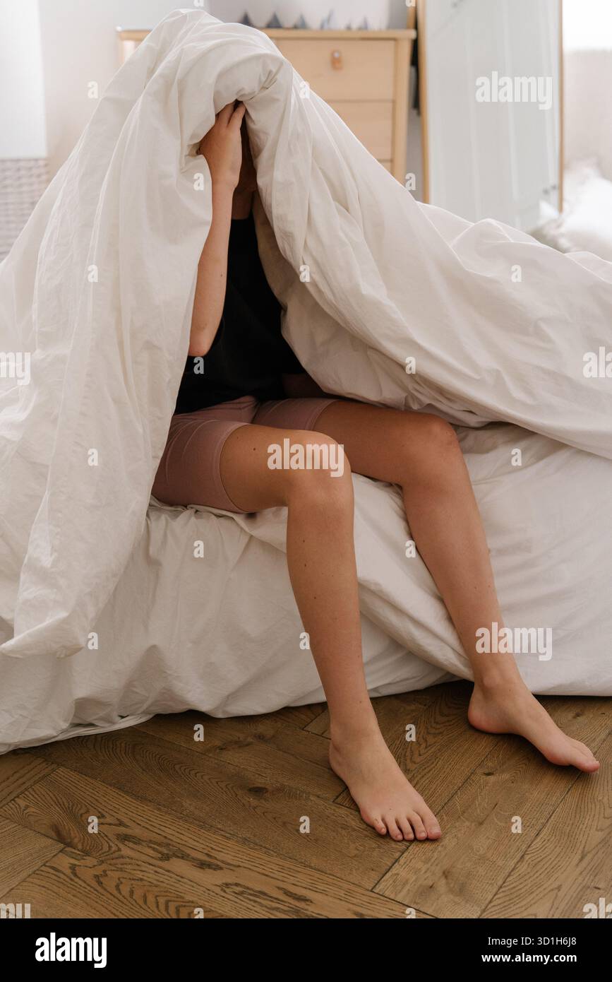 Adolescente assise sur le lit se cachant sous une couverture blanche dans la lumière du matin confortable. Banque D'Images