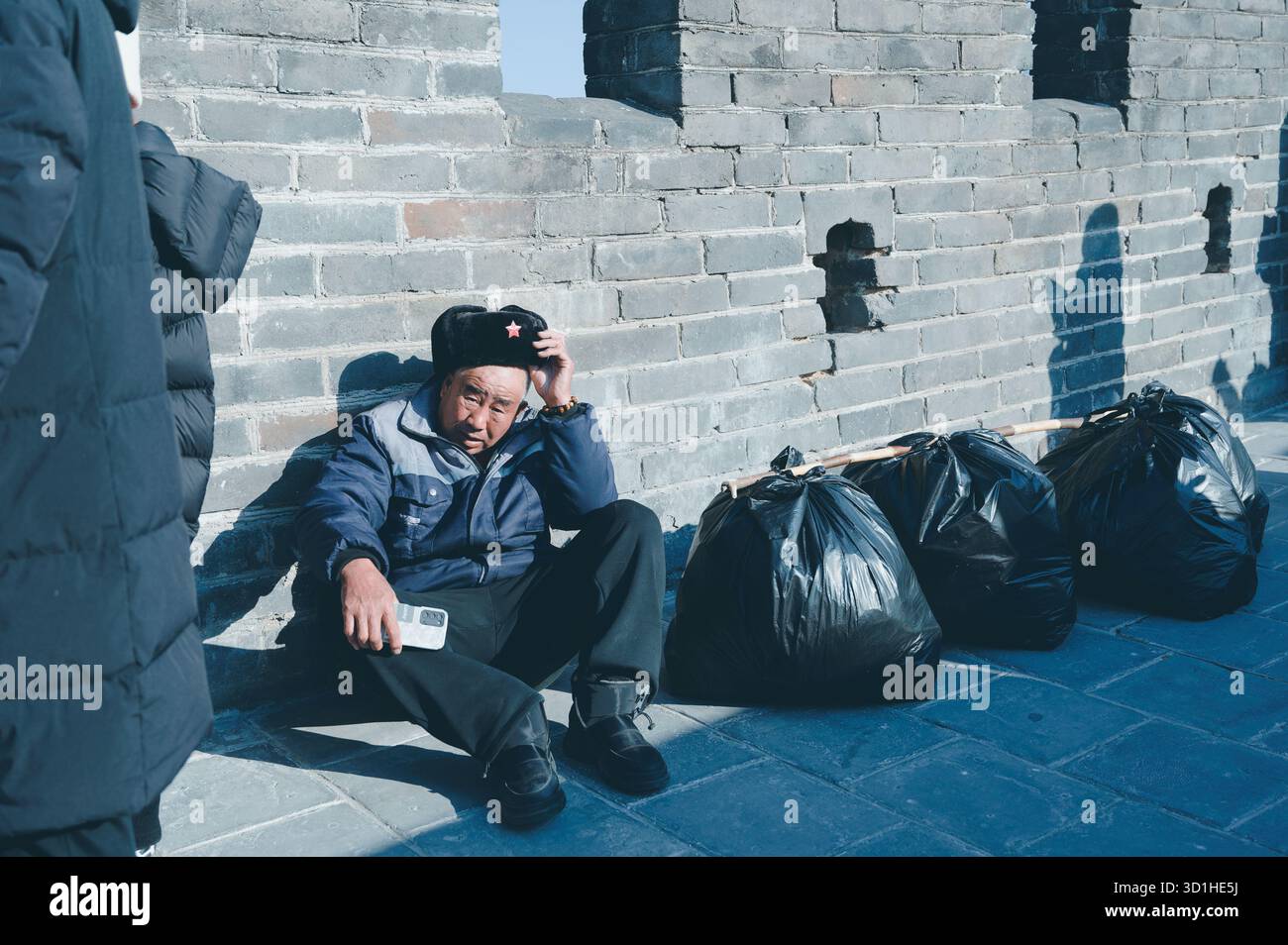 Travailleur repose contre l'ancien mur de briques de la Grande Muraille à côté de sacs poubelle soigneusement attachés sous le soleil d'hiver Banque D'Images