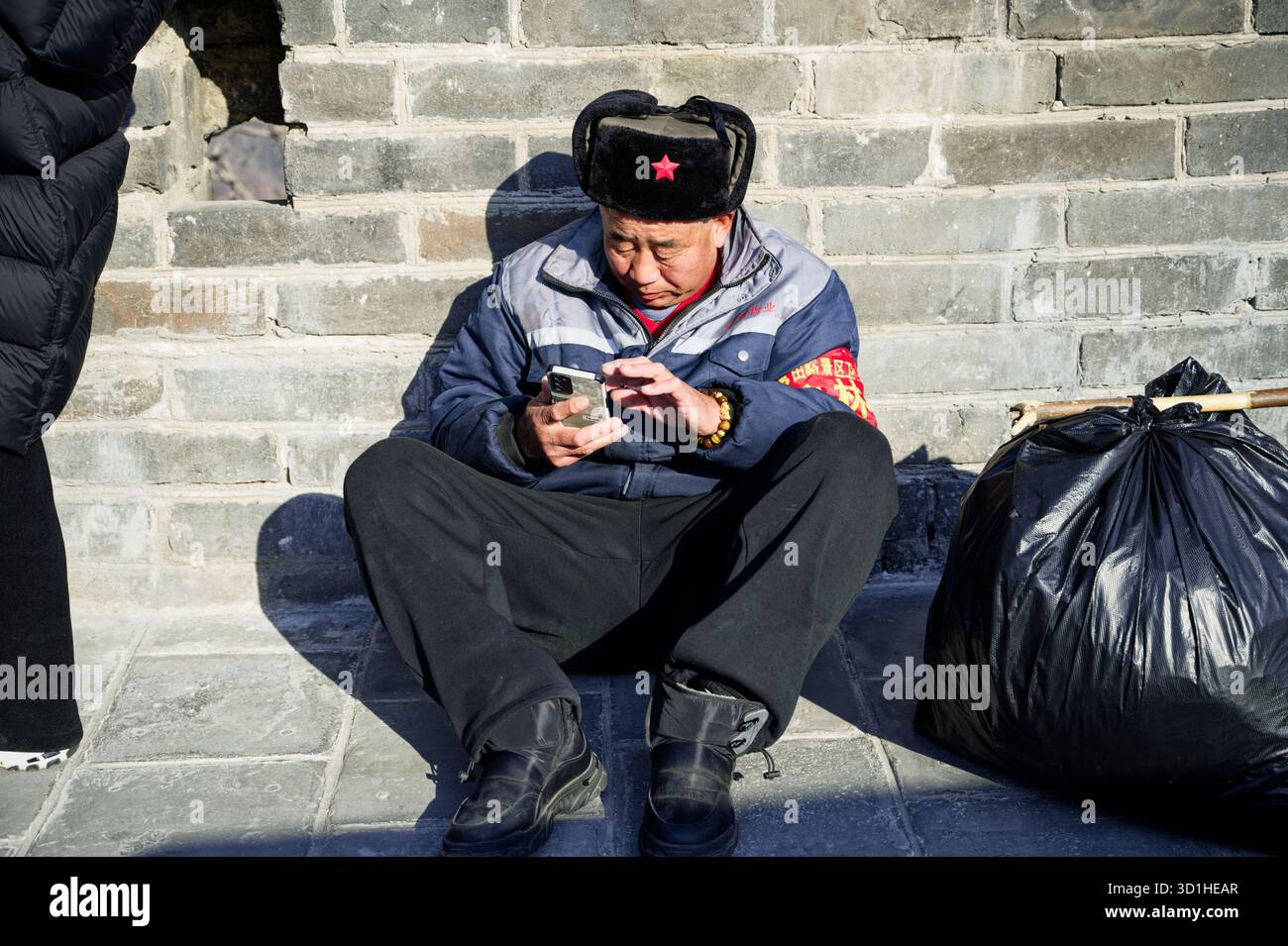 Un travailleur chinois en uniforme d'hiver est assis contre la Grande Muraille à côté de sacs poubelle soigneusement attachés Banque D'Images