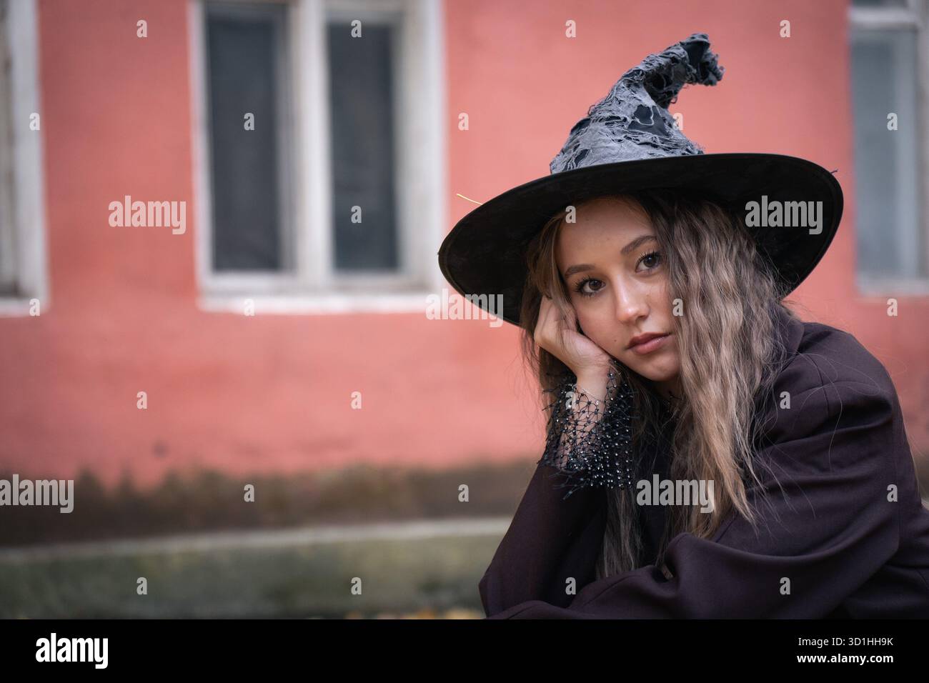 Teen Girl dans un chapeau de sorcière pose soigneusement contre un vieux bâtiment rose, mélangeant une atmosphère d'Halloween sombre avec le charme urbain Banque D'Images