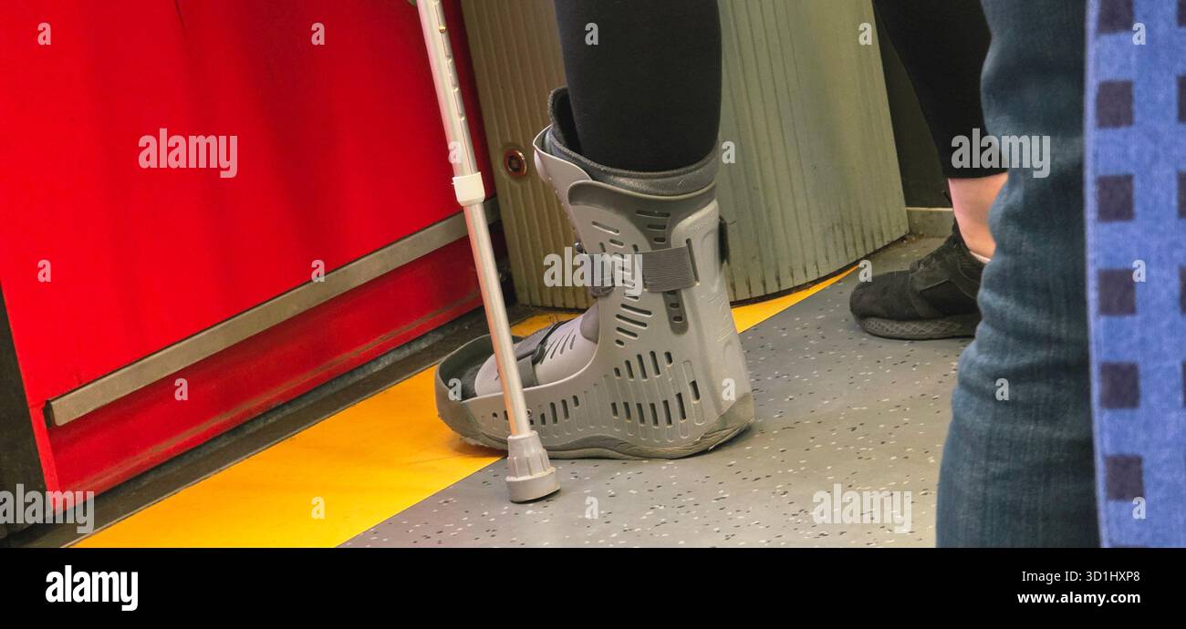 Individu portant une botte de marche et utilisant une canne, debout dans un environnement de transport public, mettant en valeur l'aide à la mobilité et la vie urbaine. Banque D'Images
