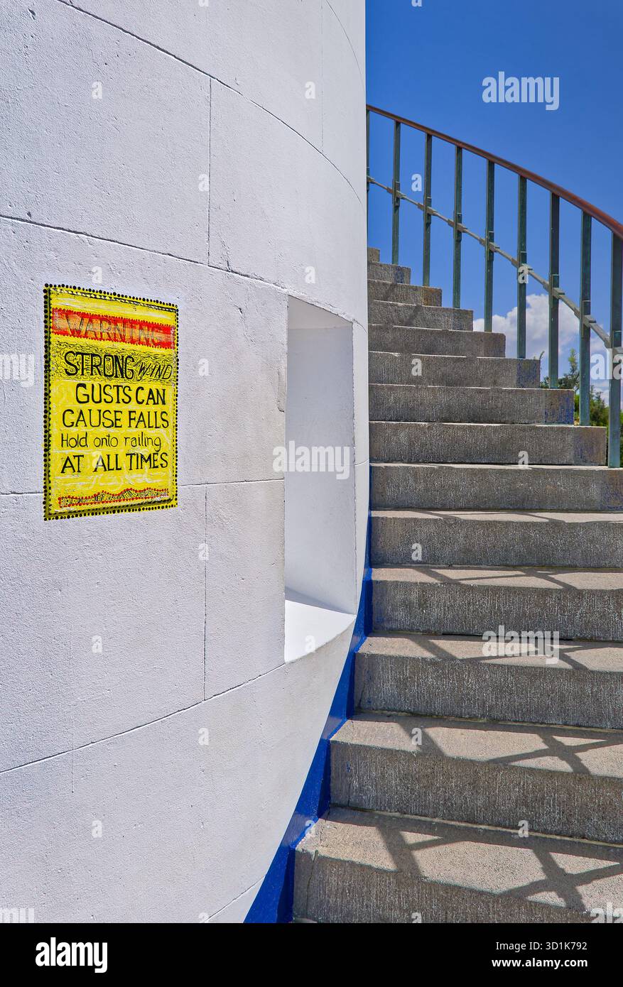 Panneau jaune avertissant de rafales de vent dangereuses sur un mur de briques blanches avec escalier en béton montant au phare de Sugarloaf point, NSW, Australie Banque D'Images