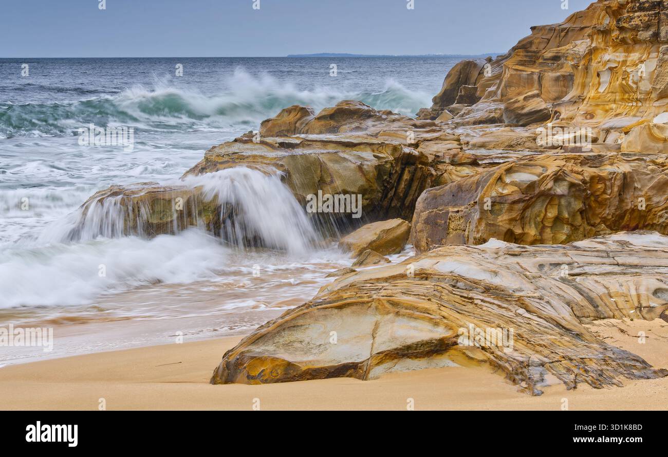 Vague d'eau floue se brisant sur des roches de grès orange et jaune par jour couvert à Maitland Bay, parc national de Bouddi, Nouvelle-Galles du Sud, Australie Banque D'Images