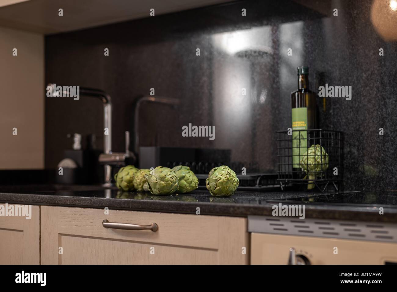 Un comptoir de cuisine moderne avec une surface en granit noir. Des artichauts verts frais sont disposés à côté d'une bouteille d'huile d'olive dans un panier en fil de fer. Banque D'Images